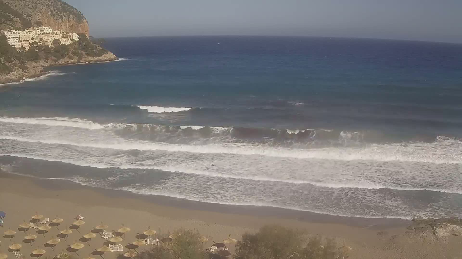 A sandy beach with numerous umbrellas and a few people is bordered by a calm sea with moderate waves breaking on the shore, backed by a rocky cliff with buildings.