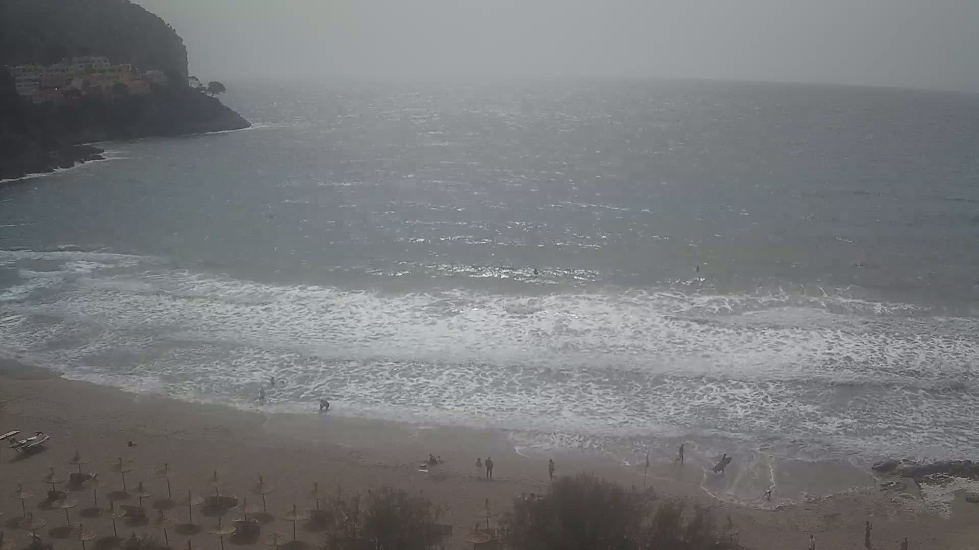 A hazy, overcast day shows a beach with small waves lapping at the shore, a few people are on the beach near closed umbrellas, and a rocky cliff is visible in the distance.