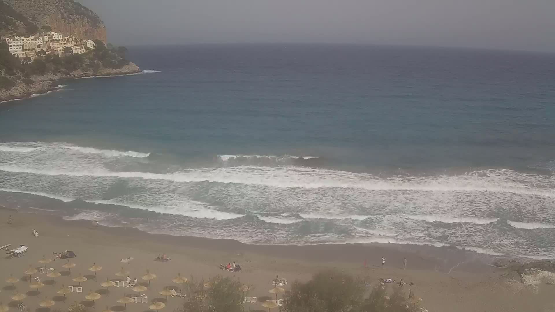 A sandy beach with small waves rolling onto the shore under an overcast sky, with people scattered on the beach and umbrellas set up.