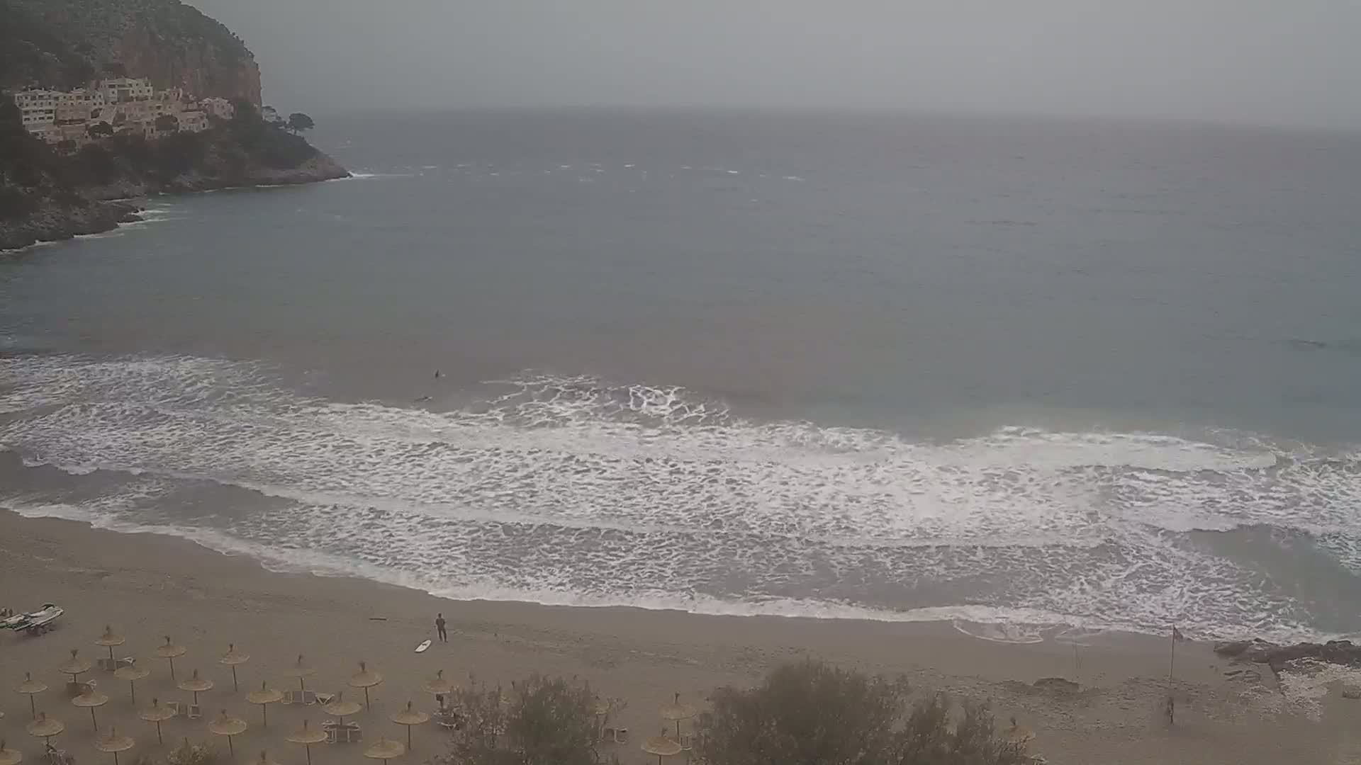 A sandy beach with small waves rolling onto the shore under an overcast sky, with a few people and numerous umbrellas visible.
