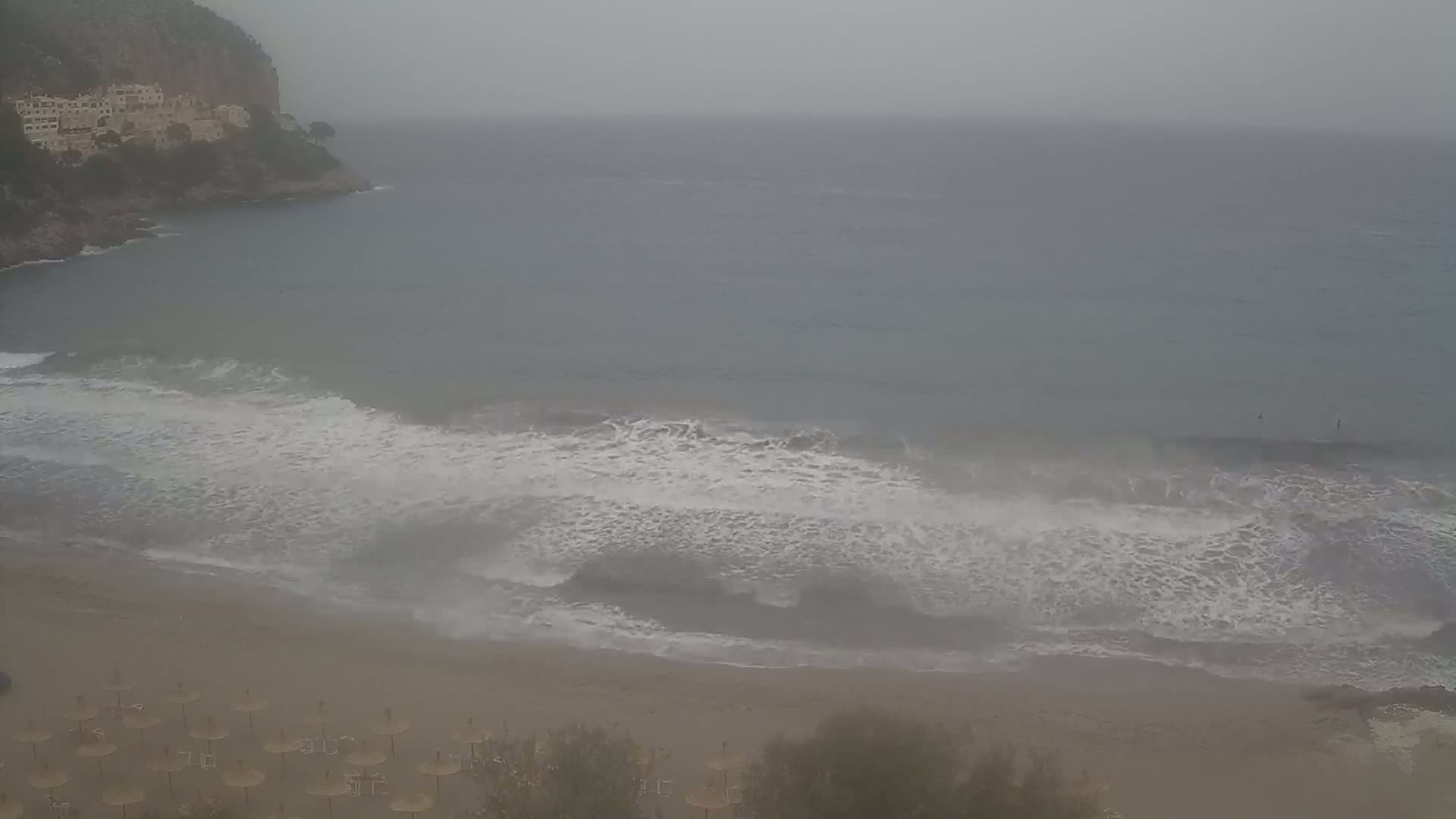 A hazy, overcast day shows moderate waves breaking on a sandy beach below a rocky cliff with buildings on top.