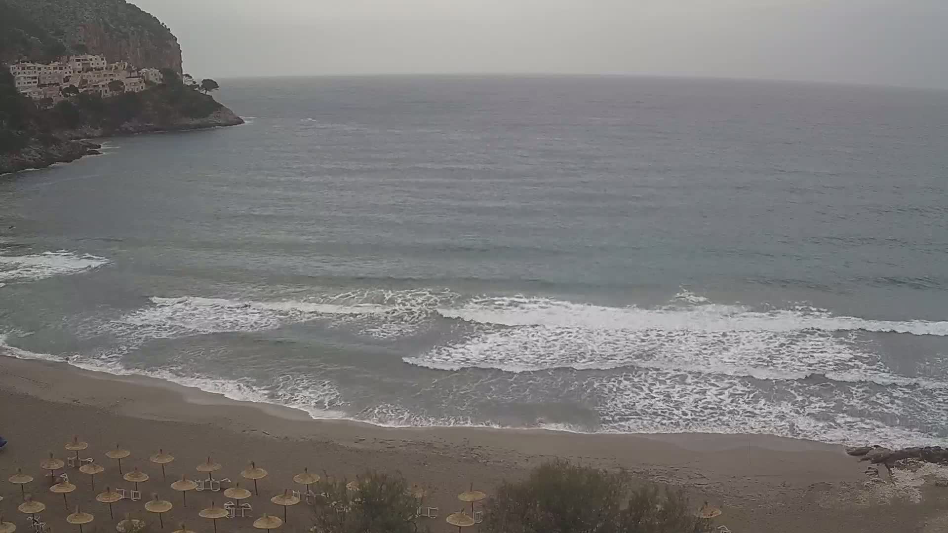 A sandy beach with numerous umbrellas is seen next to a calm sea with small waves lapping the shore, under an overcast sky.