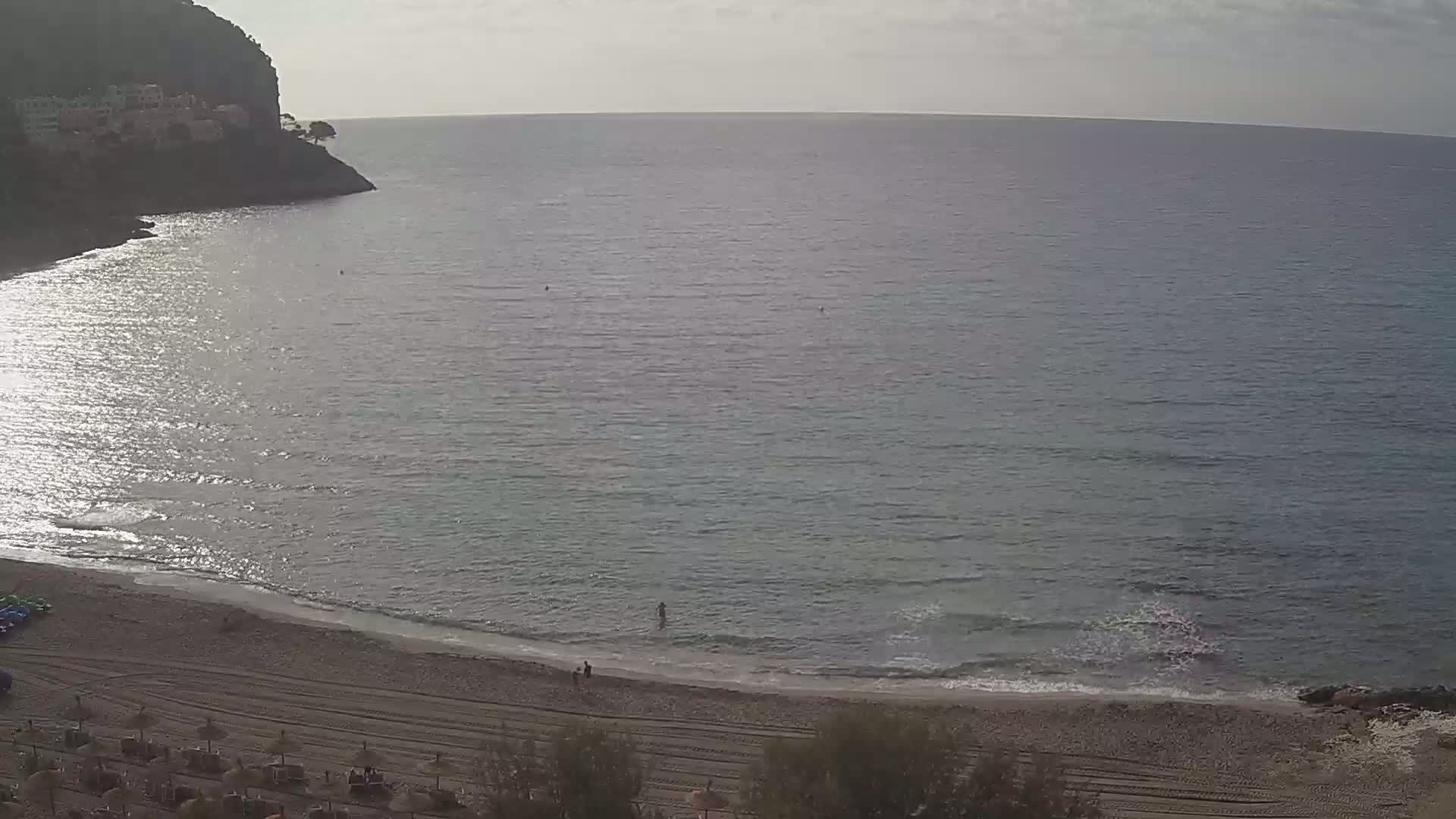 A sandy beach curves around a calm, gray-blue sea under a mostly cloudy sky, with a rocky cliff and sparse vegetation visible in the background.