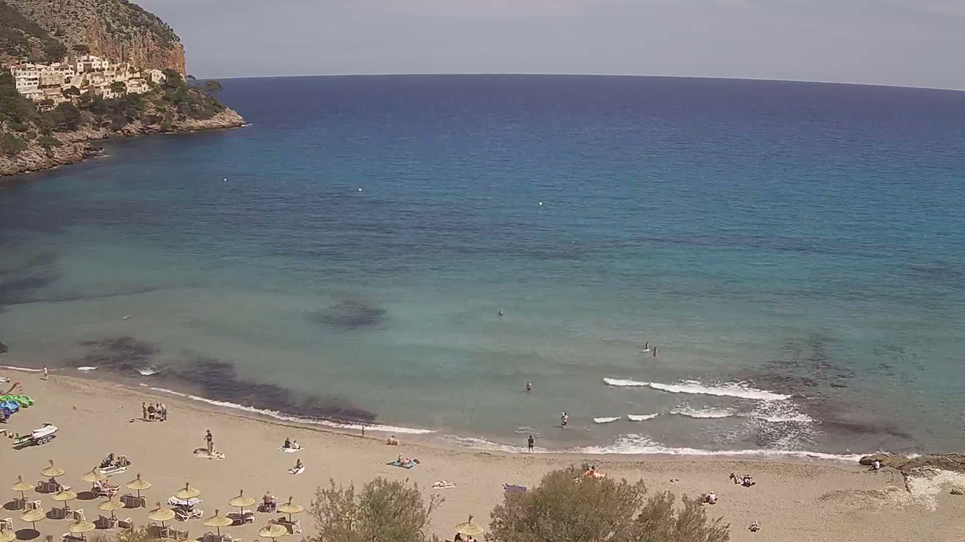 A sandy beach with scattered sunbathers and umbrellas sits below a rocky cliff overlooking a calm, clear turquoise sea on a sunny day.