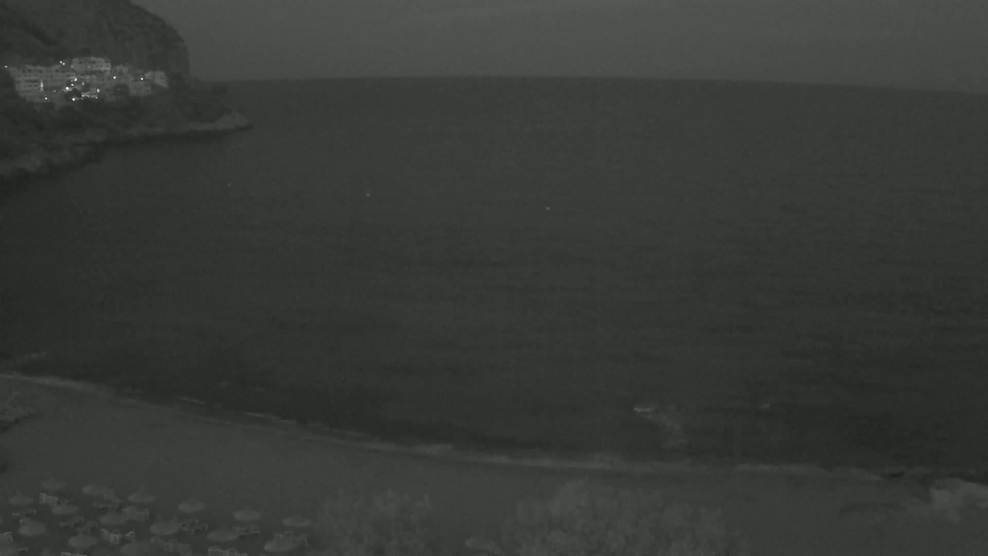A dark, calm sea laps a sandy beach at the foot of a dimly lit cliff at night.