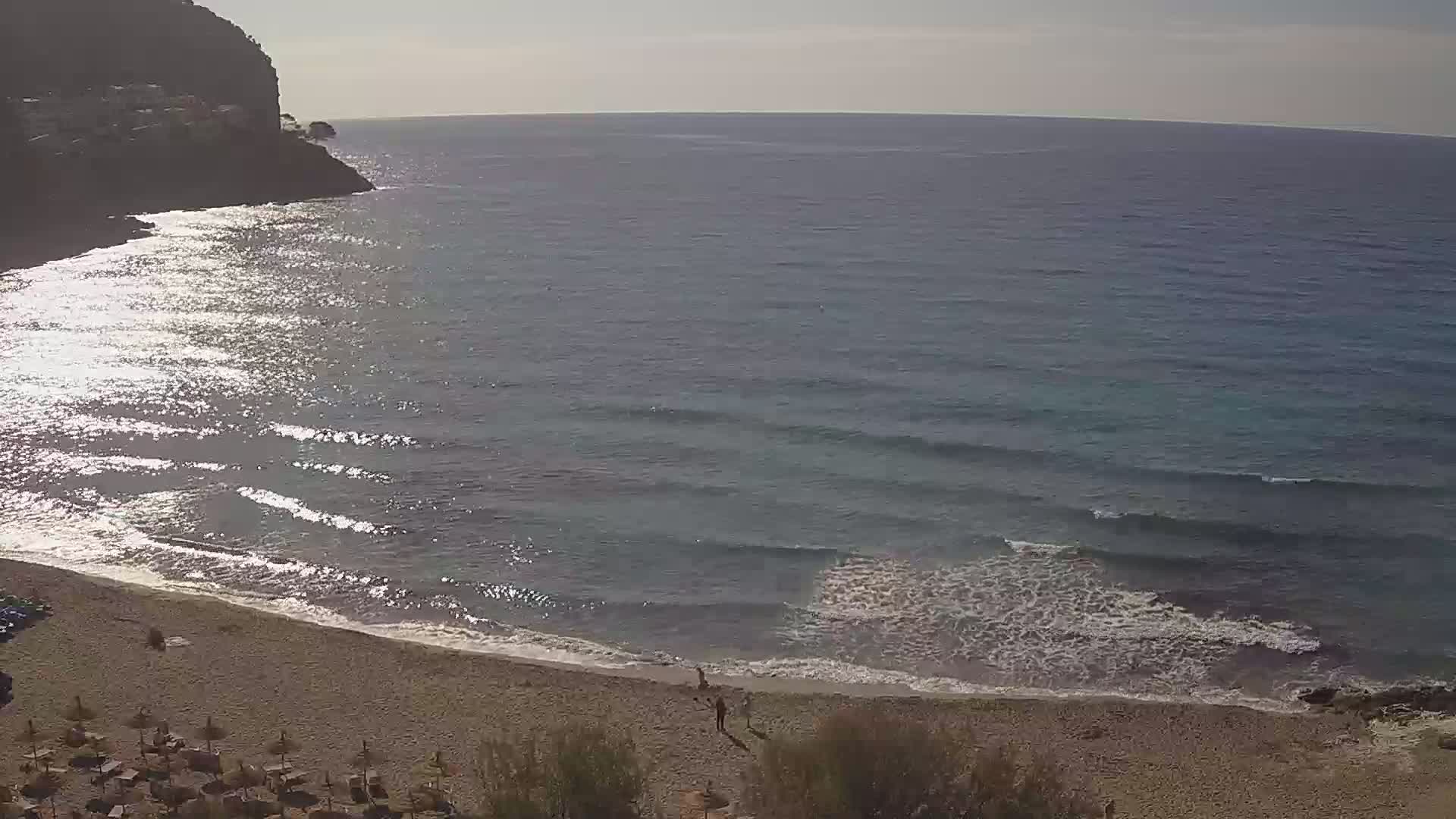 A sandy beach with calm, gently lapping waves meets a dark blue sea, backed by a rocky cliff face under a bright, sunny sky.