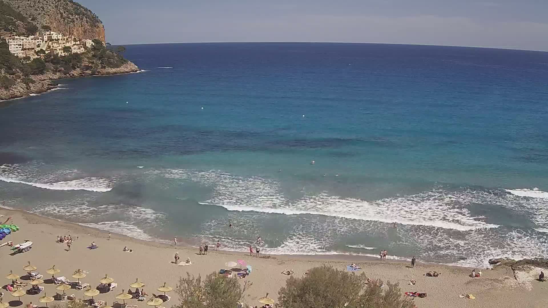 A sandy beach with people sunbathing and swimming in calm, turquoise water, backed by a rocky cliff with buildings on top, under a clear, sunny sky.