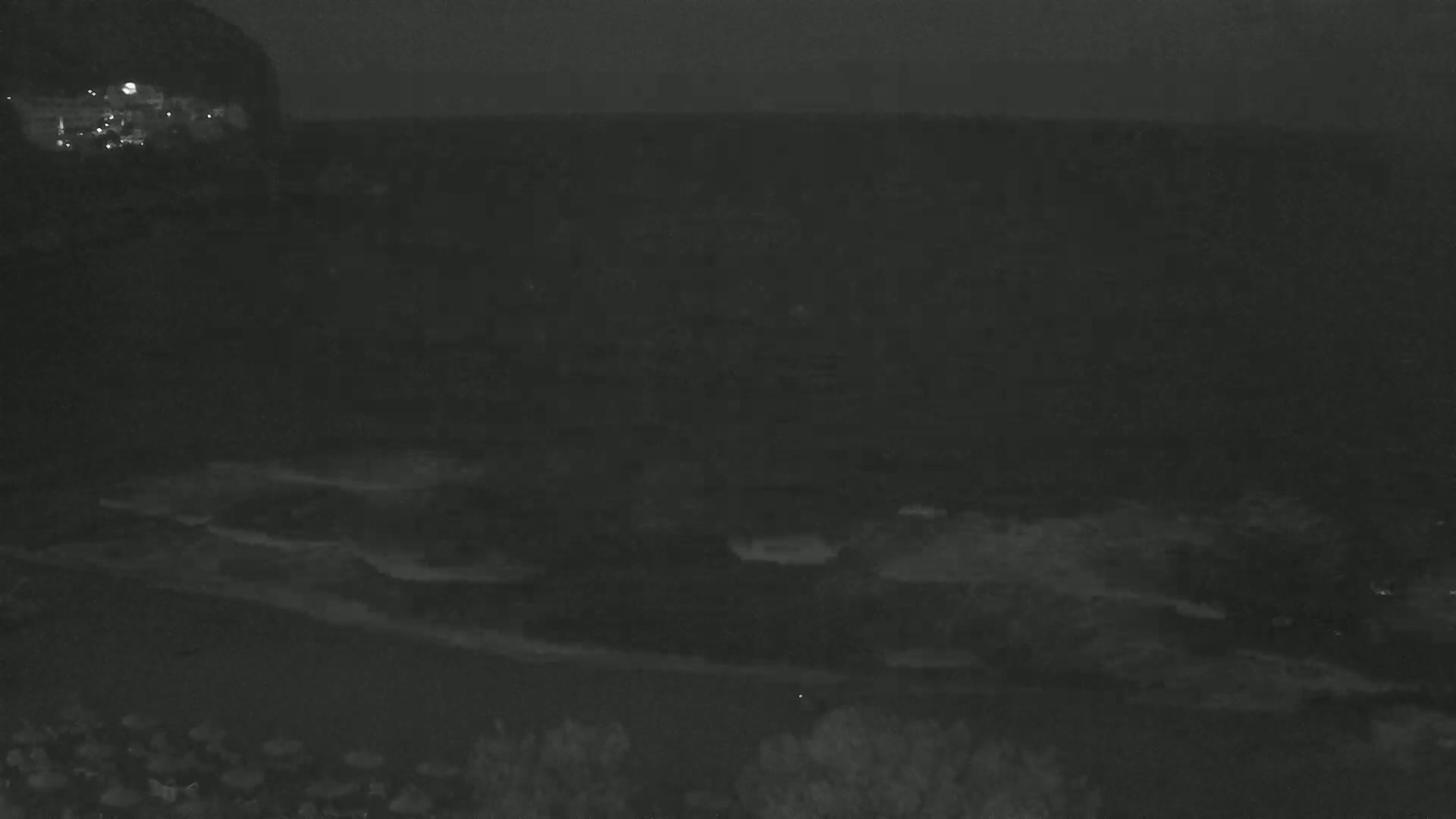 A dark, nighttime view of a beach with calm waves lapping the shore, under a mostly clear sky.