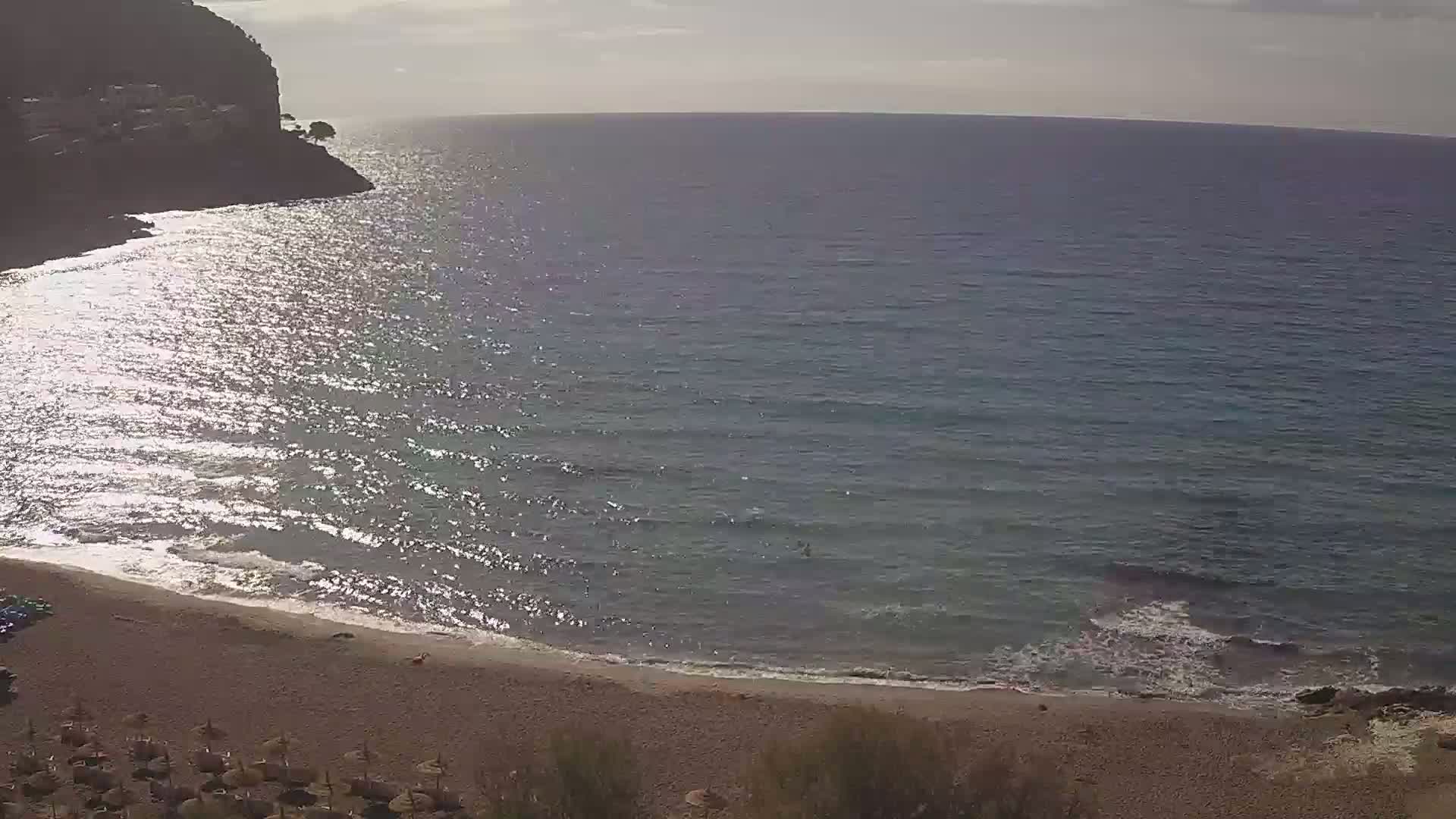 A sandy beach curves around a calm, sunlit sea next to a rocky cliff under a mostly cloudy sky.