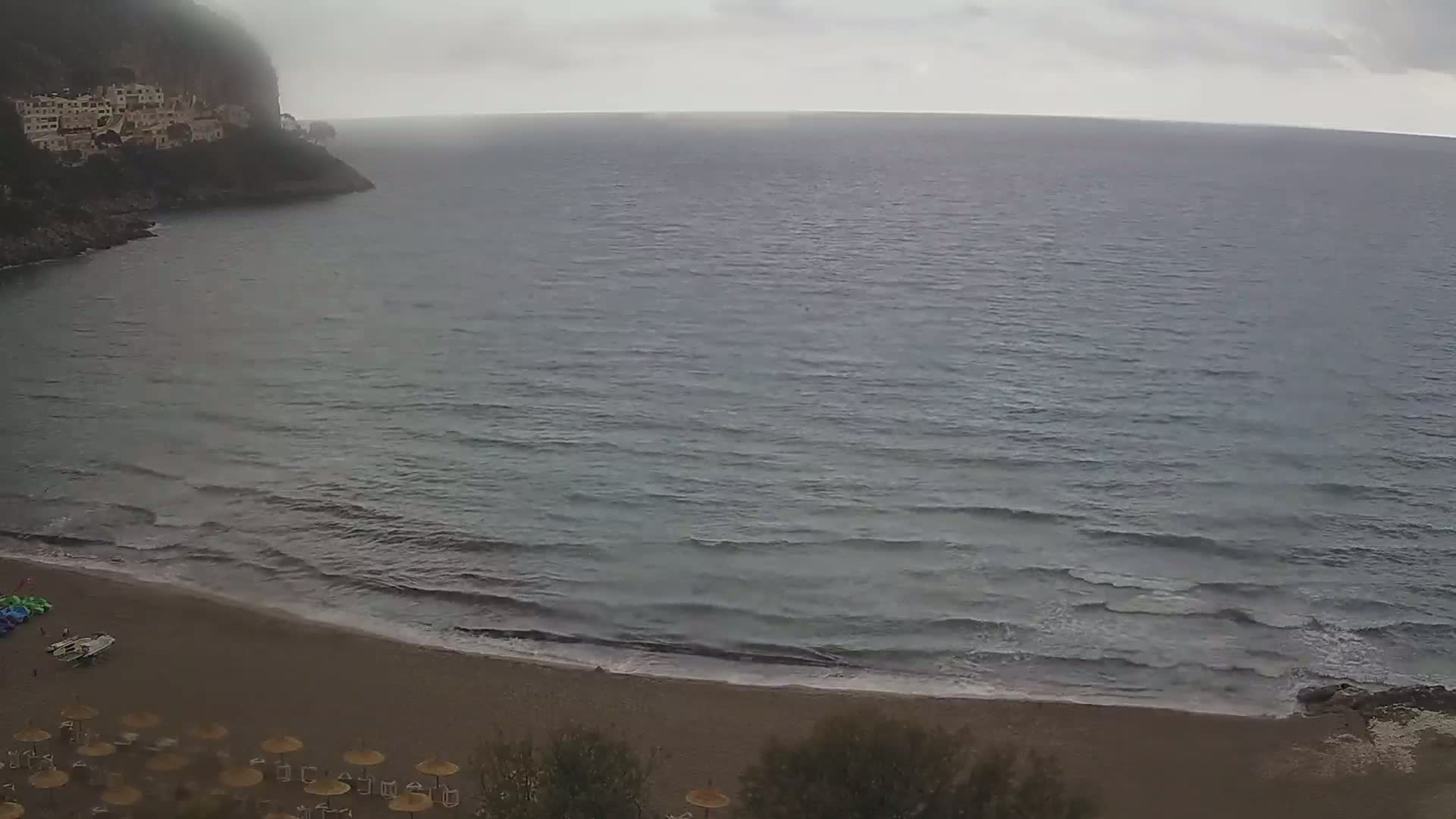A sandy beach with small waves and a few closed umbrellas sits below a cliff with buildings on a cloudy day.