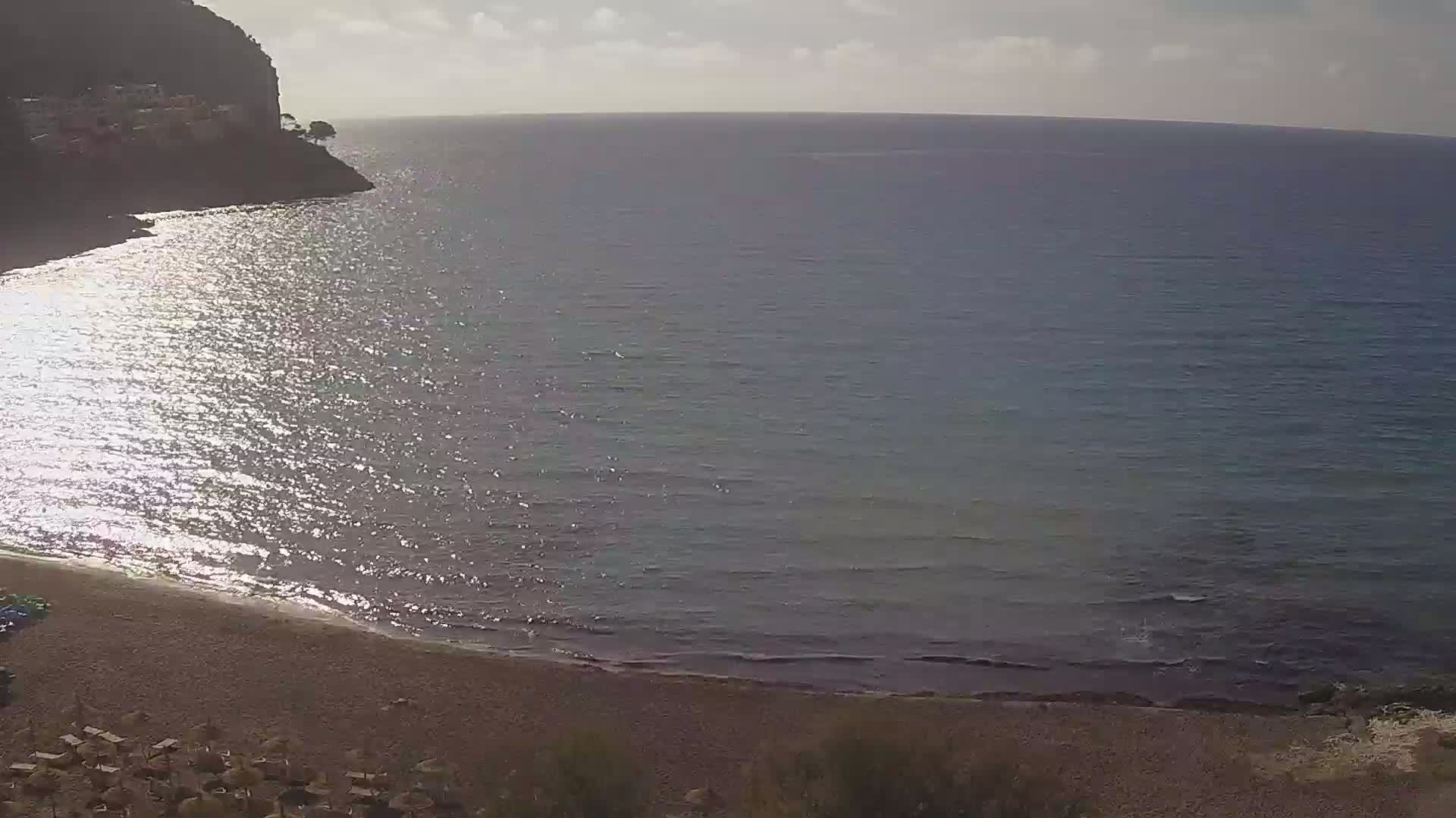 A sandy beach curves along a calm, sparkling sea next to a rocky cliff under a partly cloudy sky.