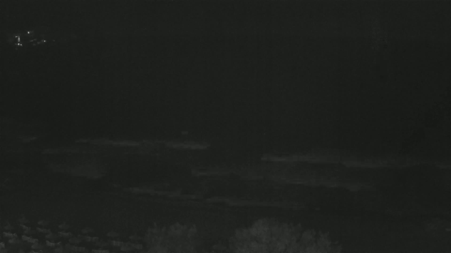A dark, nighttime scene shows a low horizon line with faint, indistinct shapes of vegetation and a few distant lights in the upper left.