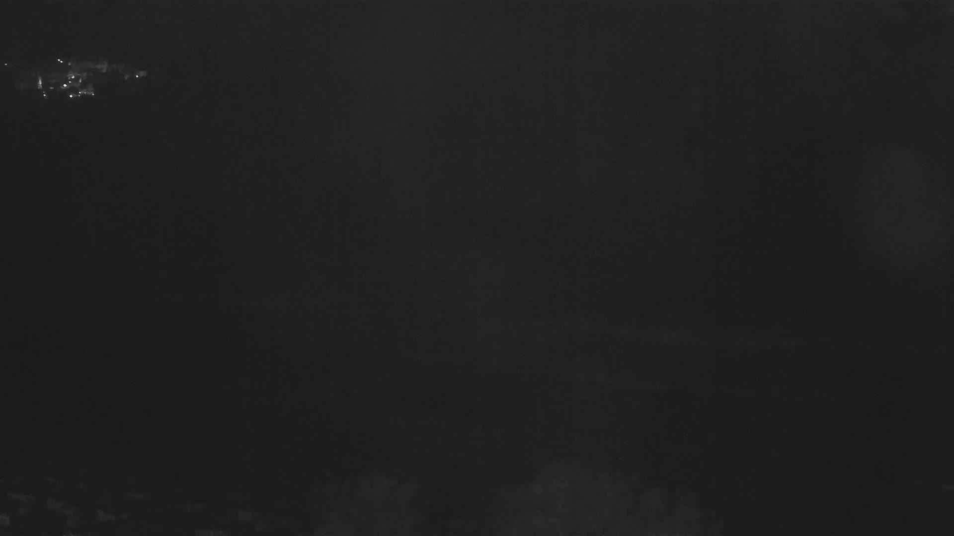 A dark, possibly nighttime, image shows a very dimly lit, indistinct landscape with a few small, faint lights in the distance.