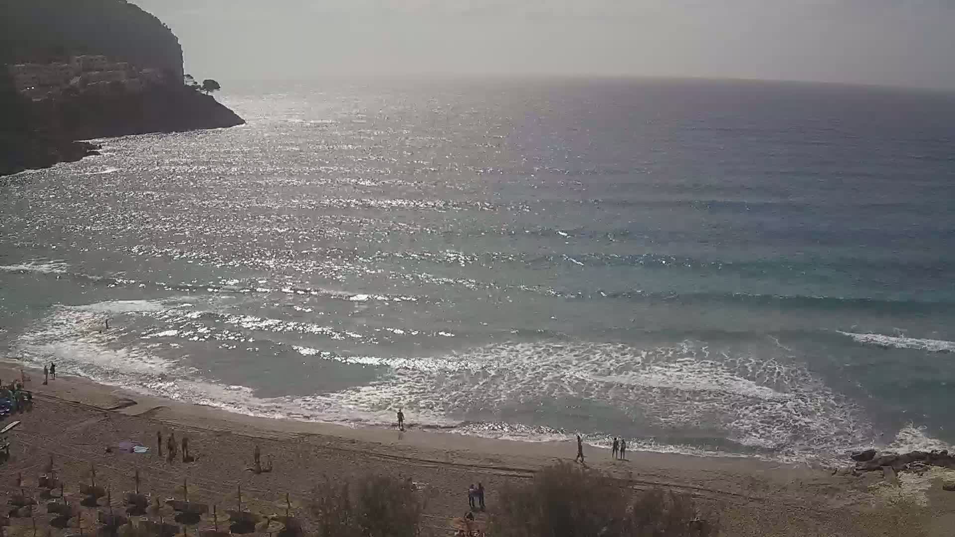 A sandy beach with a few people on it is bordered by a rocky cliff, and a somewhat choppy sea extends to the horizon under a hazy sky.