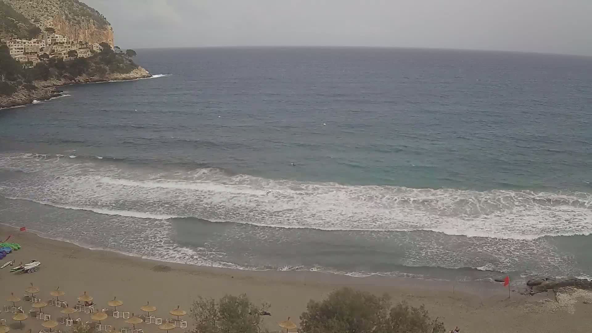 A sandy beach with moderately sized waves rolling onto the shore under an overcast sky, backed by a rocky cliff with buildings.