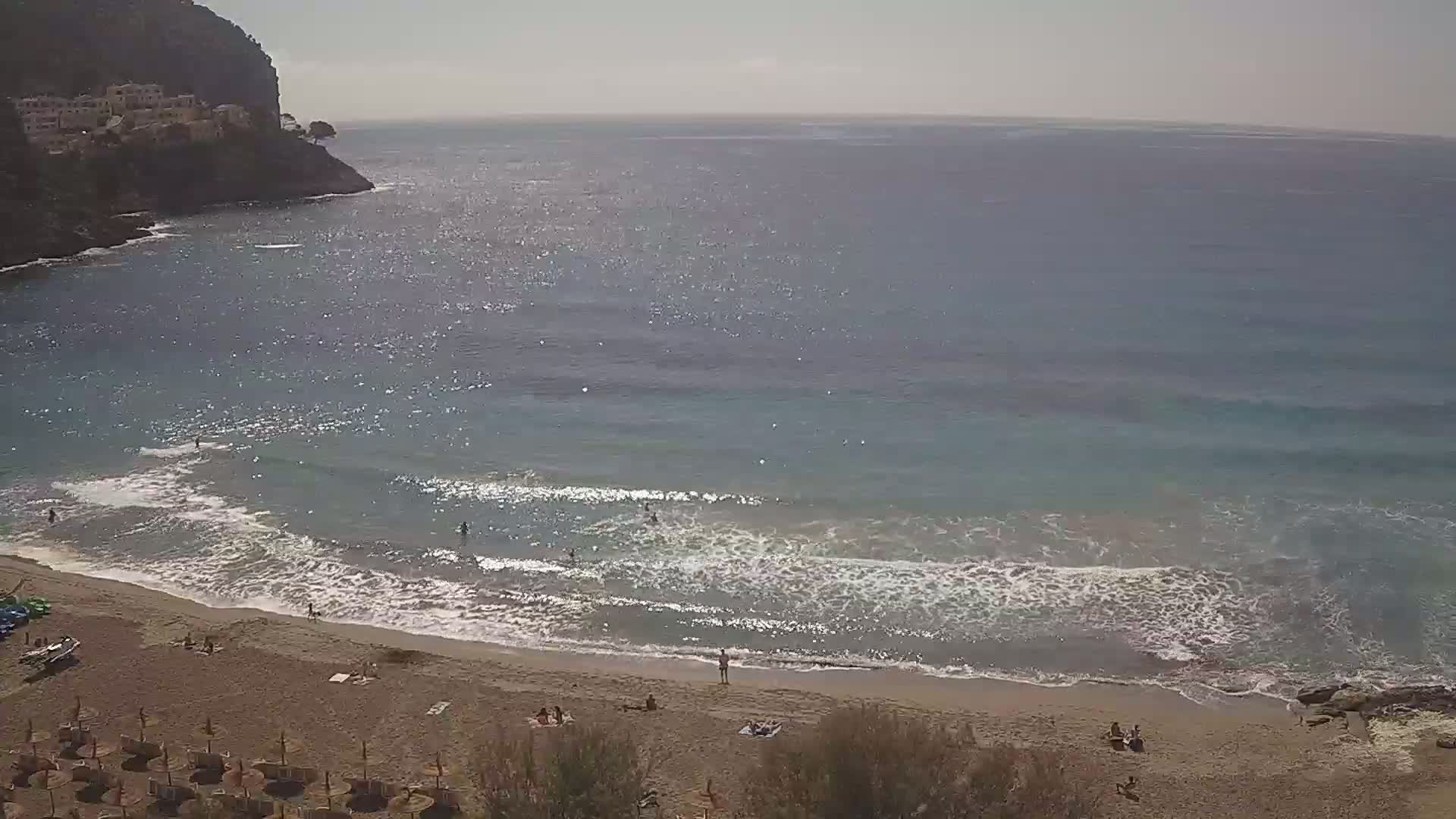 A sandy beach with small waves lapping the shore, under a sunny sky, is nestled beside a rocky cliff next to a calm sea.