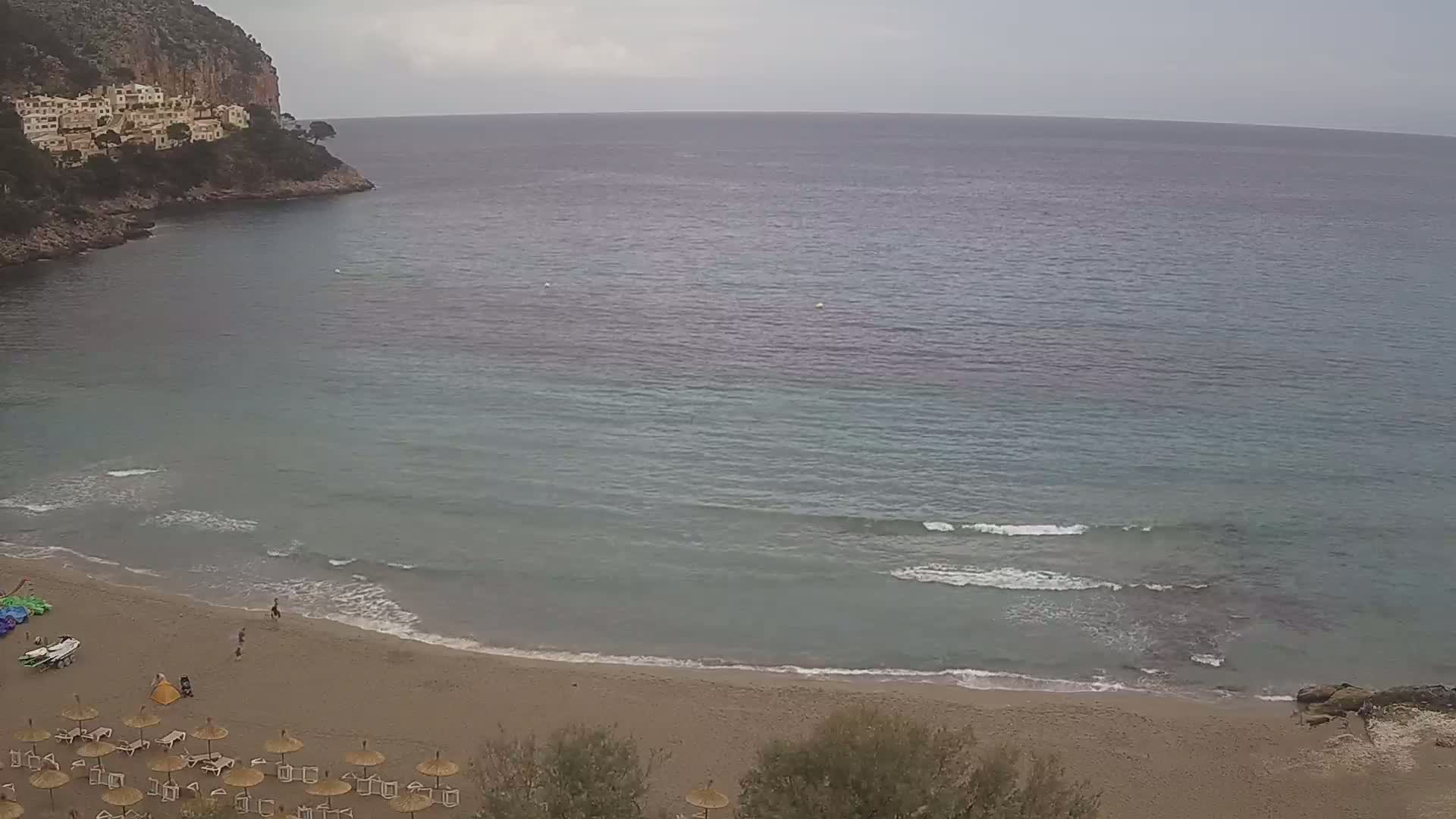 A sandy beach with calm waves and a few people on it is nestled in a cove next to a rocky cliff face under an overcast sky.