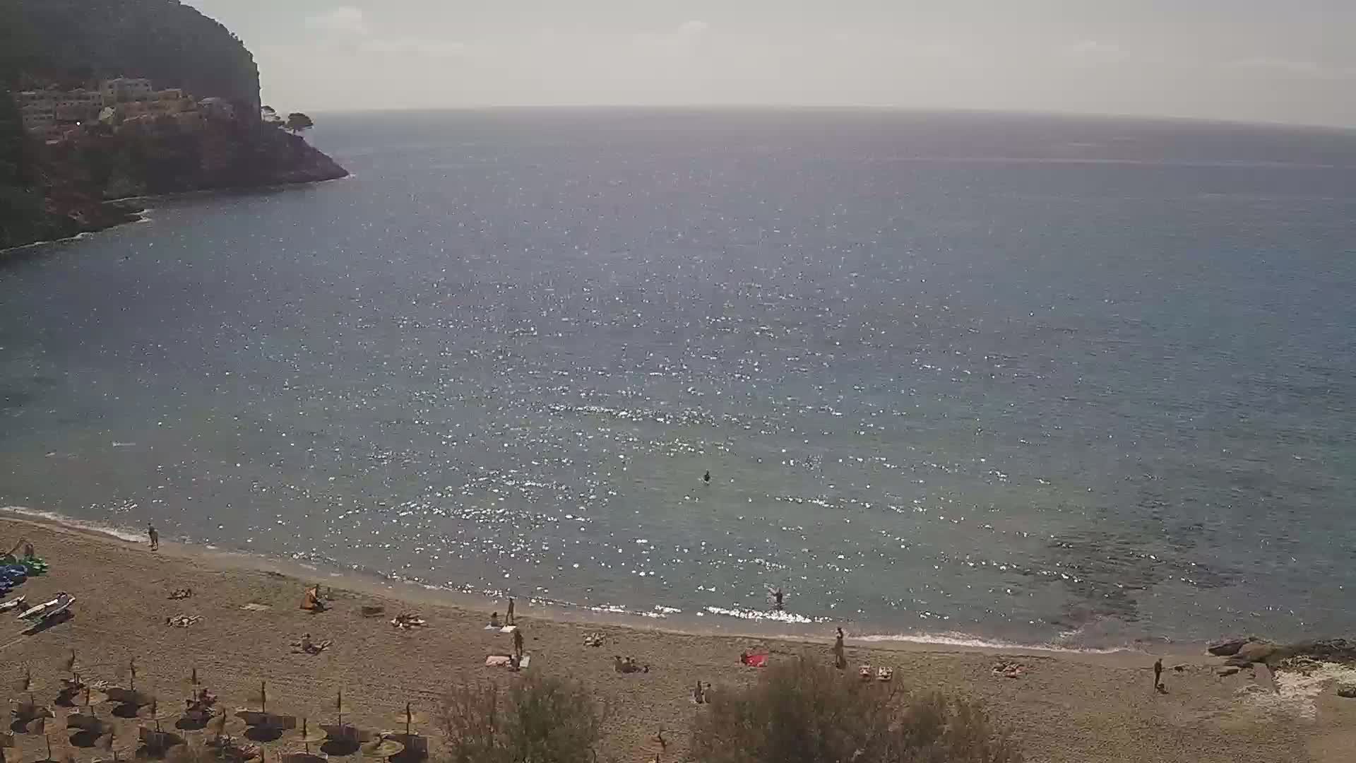 A sandy beach with scattered sunbathers and umbrellas is nestled in a cove, with a calm, glittering sea extending to a hazy horizon under a mostly clear sky.