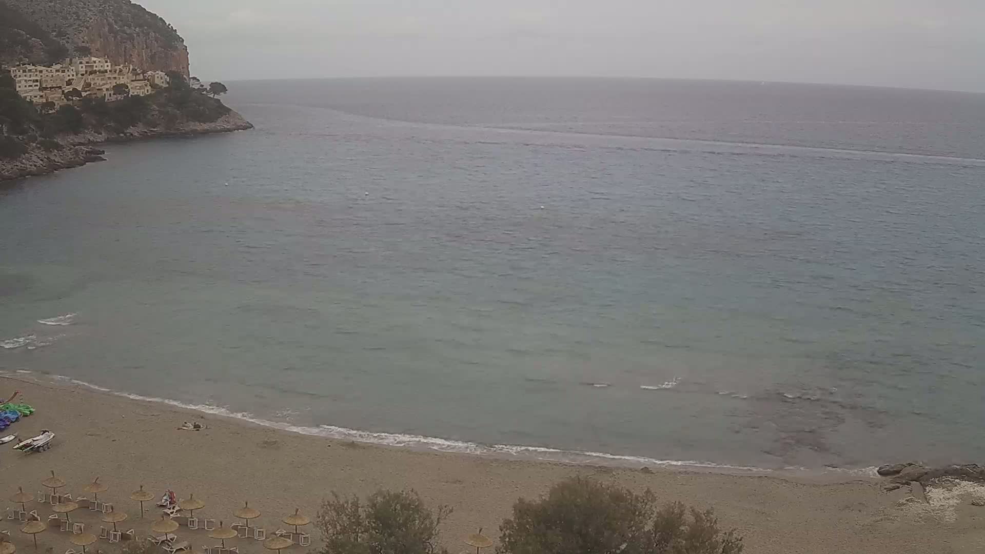 A sandy beach with a few people and numerous umbrellas sits below a rocky cliff overlooking a calm, gray sea under an overcast sky.