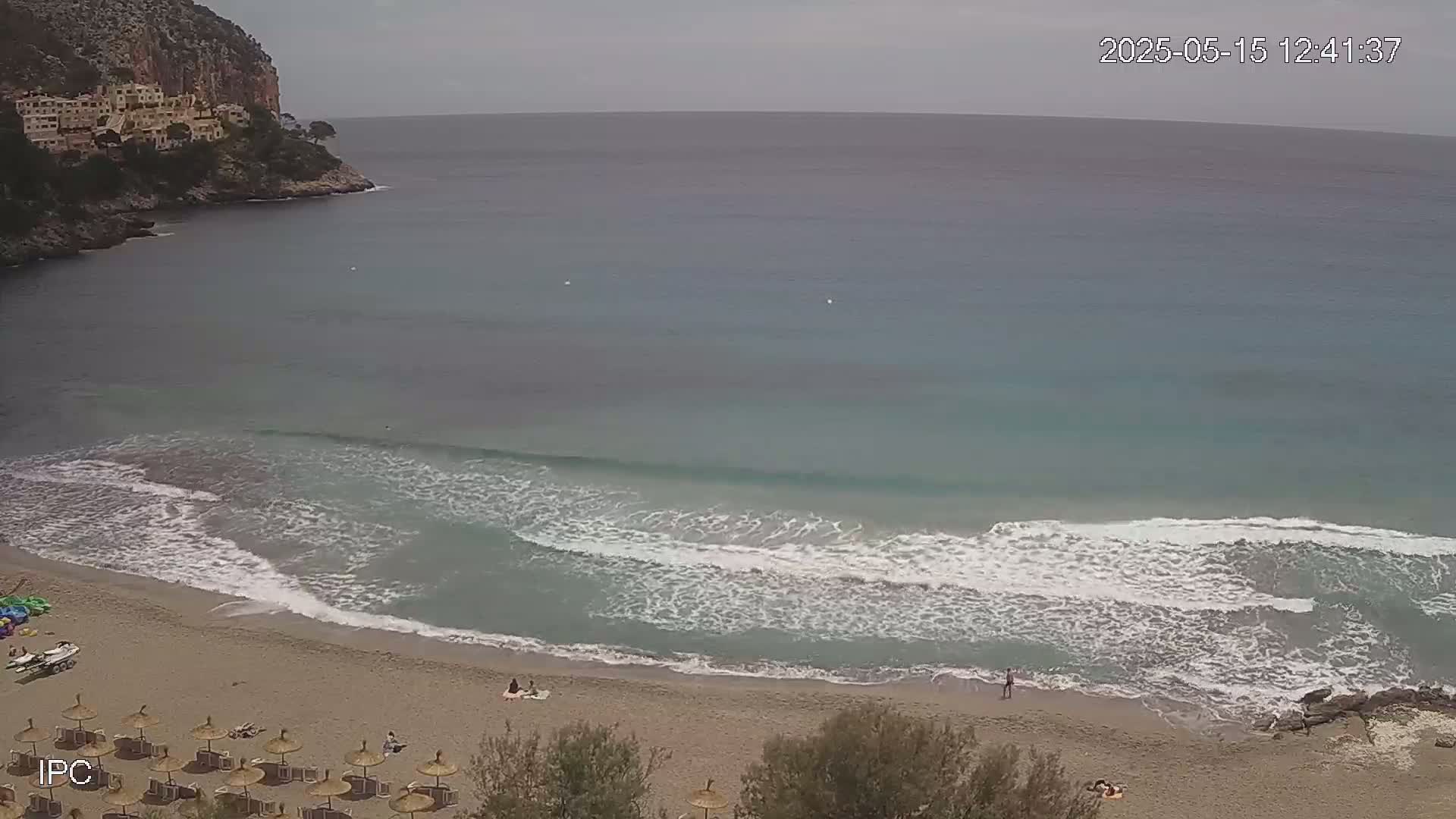 A sandy beach with small waves gently lapping the shore is nestled in a cove beneath a cliff, under an overcast sky.