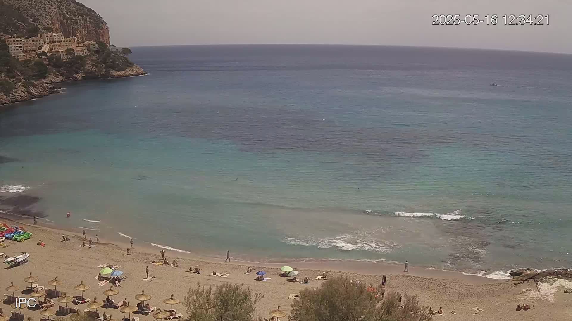 A sandy beach with people sunbathing and swimming in clear, shallow turquoise water, backed by a rocky cliff under a mostly sunny sky.