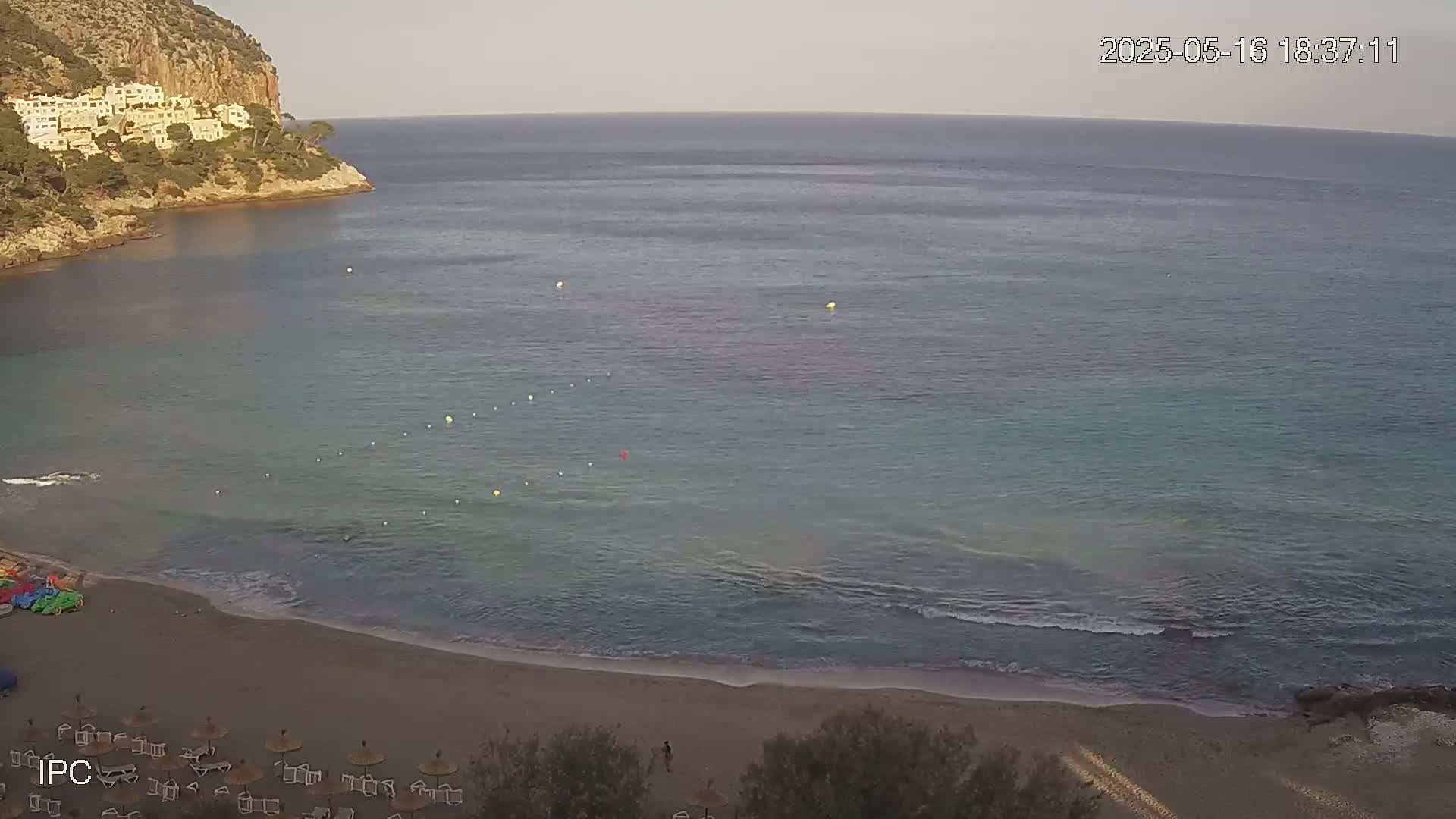 A sandy beach with calm, clear turquoise water laps gently at the shore, backed by a rocky cliff with buildings on top under a mostly clear sky.