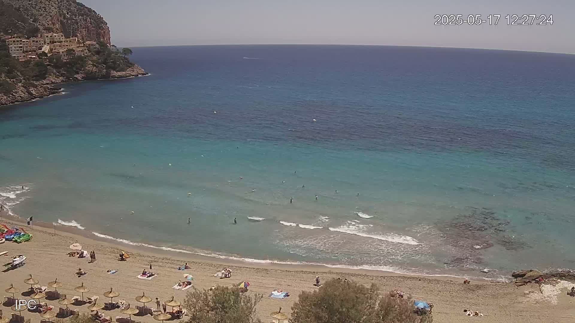 A sandy beach with turquoise water and a few people swimming and sunbathing is nestled in a cove next to a rocky cliff on a sunny day.