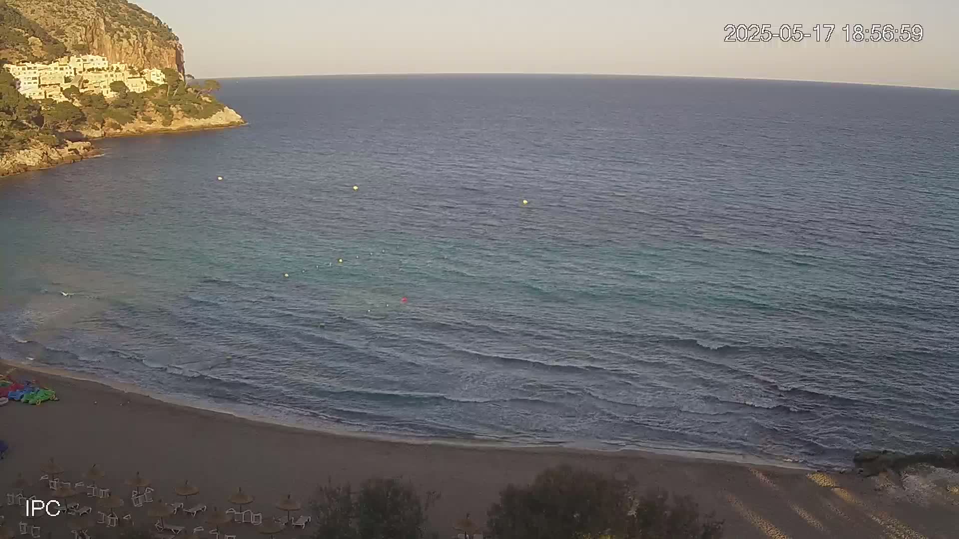 A sandy beach with calm waves laps the shore near a rocky cliff with buildings under a mostly clear evening sky.