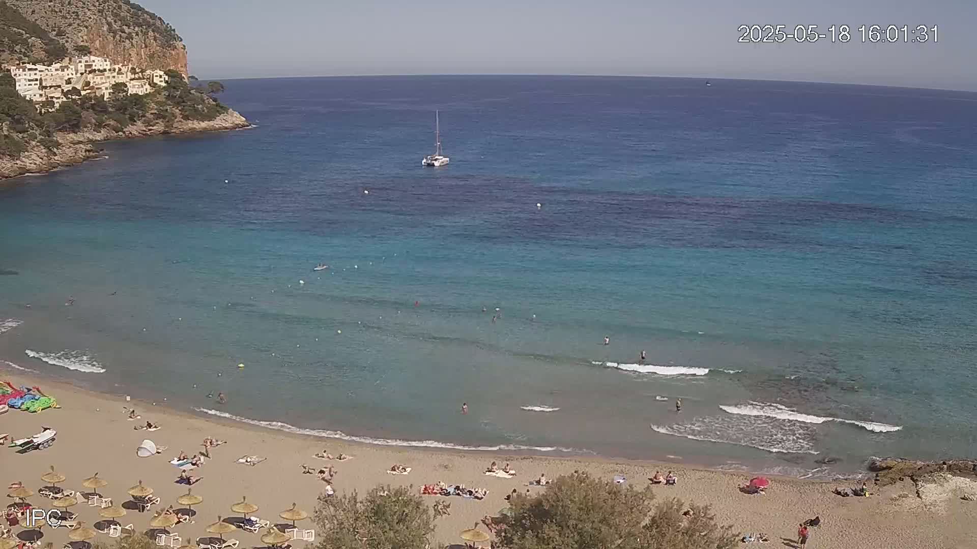 A sandy beach with scattered sunbathers and umbrellas is nestled in a cove of clear, teal water under a sunny sky, with a sailboat visible in the distance.