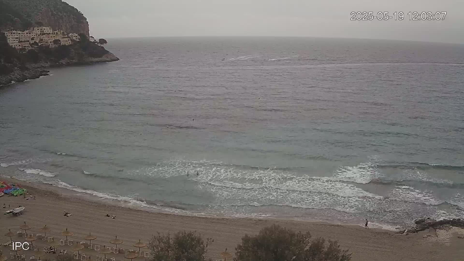 A sandy beach with small waves and a few people on it is nestled in a cove below a cliff with buildings on top, all under an overcast sky.