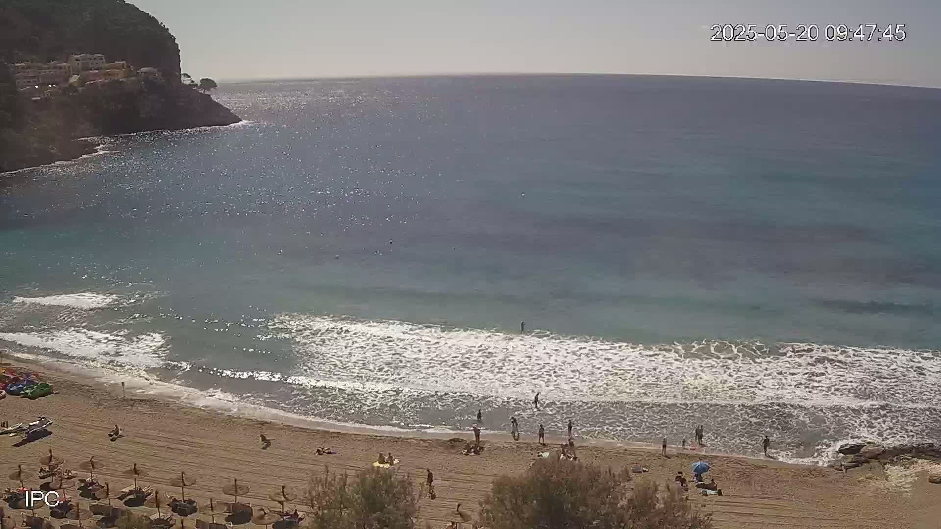 A sandy beach with people sunbathing and swimming in calm, shallow water next to a rocky cliff under a sunny sky.