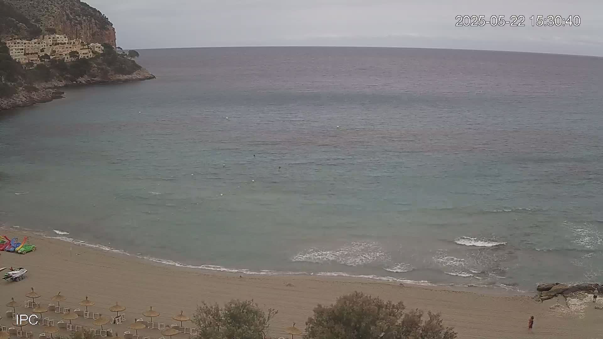 A sandy beach with calm, light-blue water and a few people is nestled in a cove next to a rocky cliff under an overcast sky.