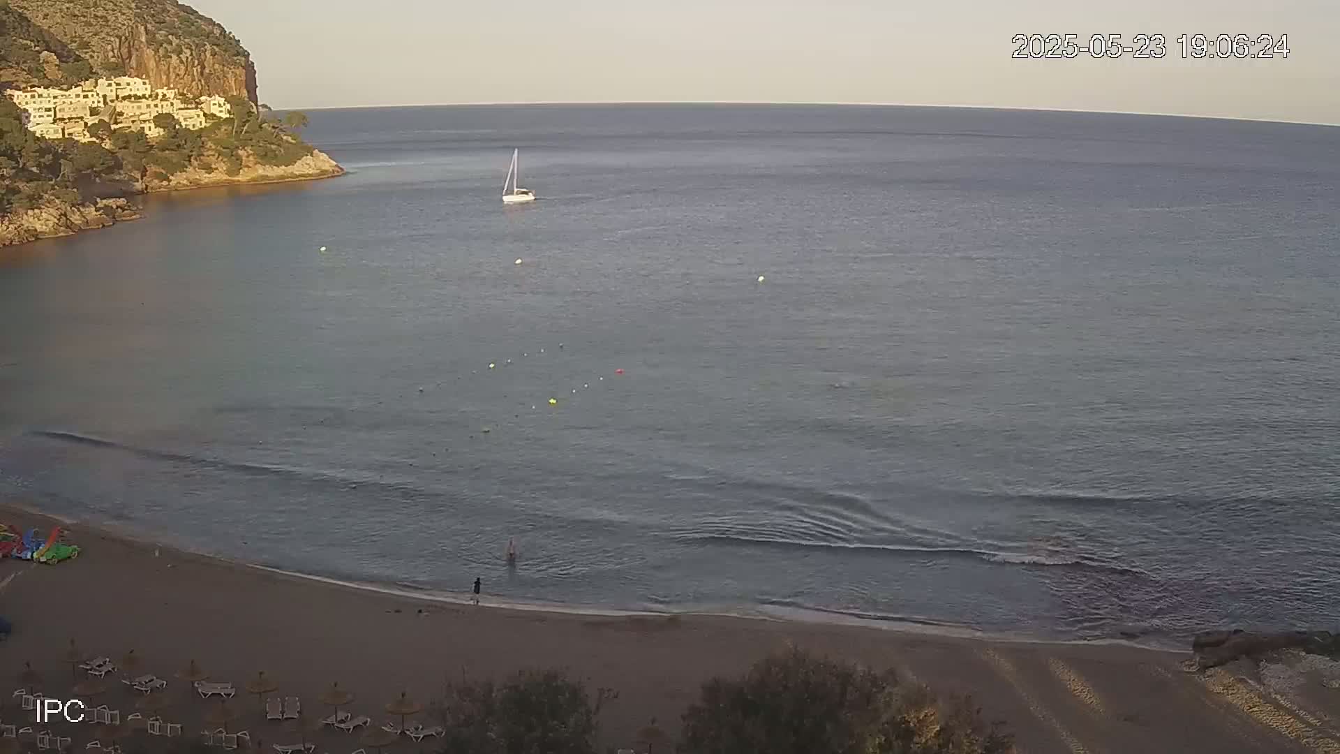 A sandy beach with calm waves and a sailboat on the water, backed by a rocky cliff and buildings.