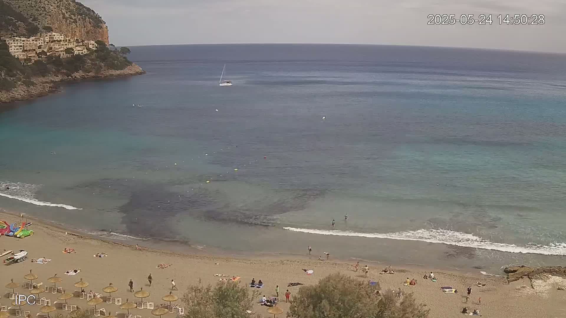 A sandy beach with scattered sunbathers and a calm, clear blue-green sea curves around a rocky headland under an overcast sky.