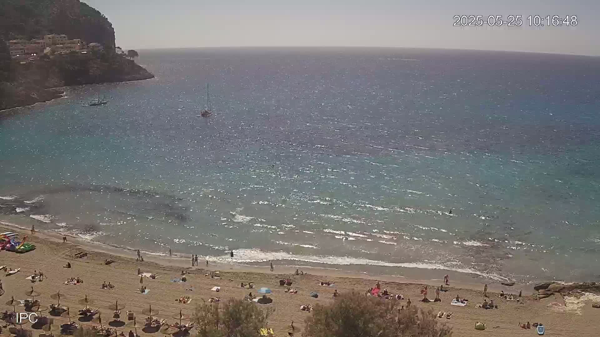 A sandy beach with people sunbathing and swimming, bordered by a calm, blue sea and a rocky cliff under a sunny sky.