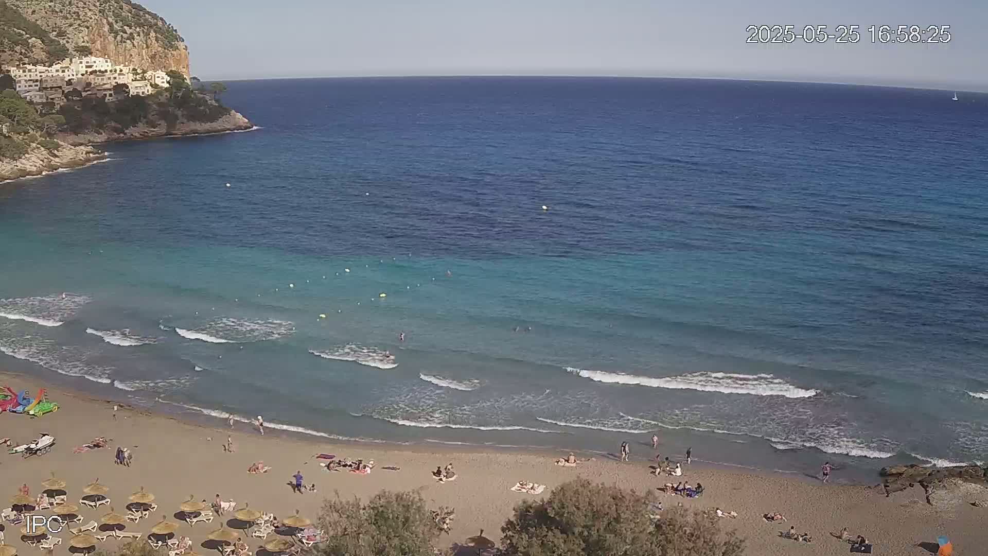 A sandy beach with scattered sunbathers and small waves laps against a clear, turquoise sea, backed by a rocky cliff with buildings on top, under a sunny sky.
