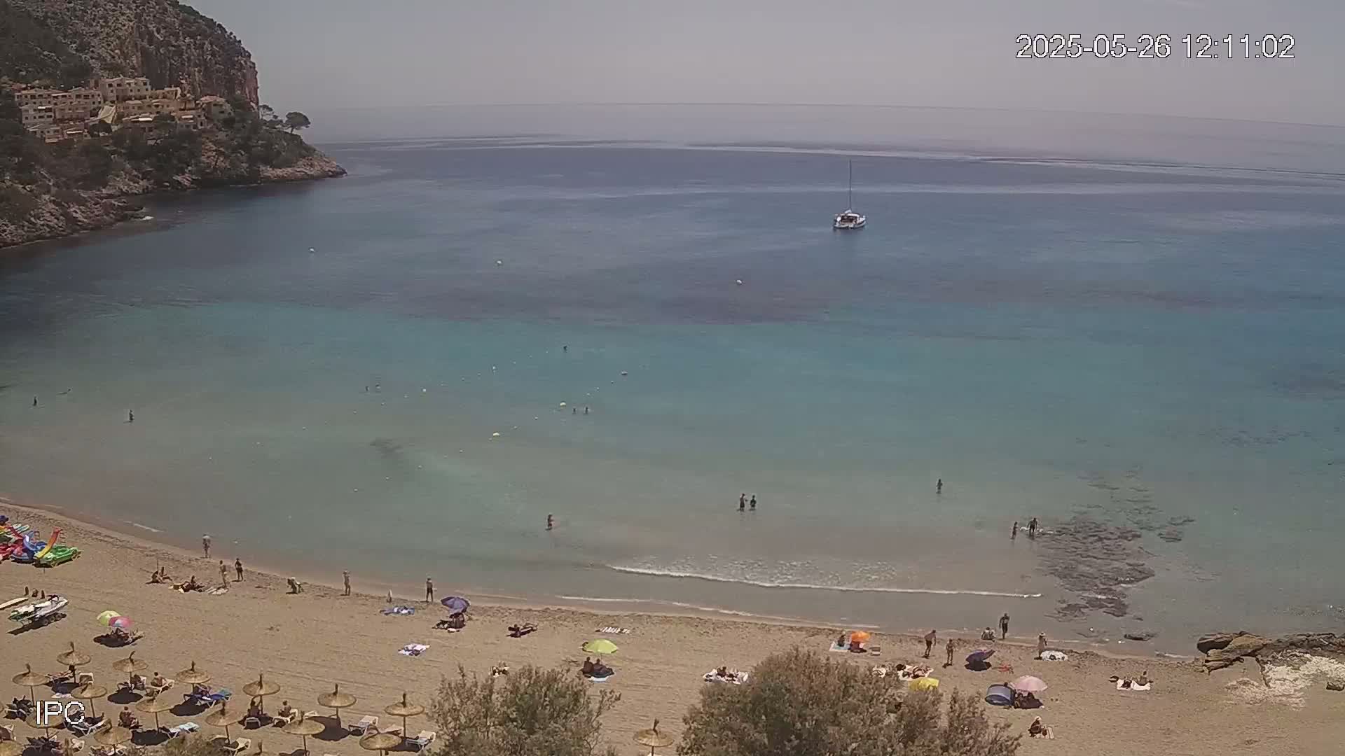 A sandy beach with bathers and umbrellas sits below a calm, clear turquoise sea next to a rocky cliff on a sunny day.