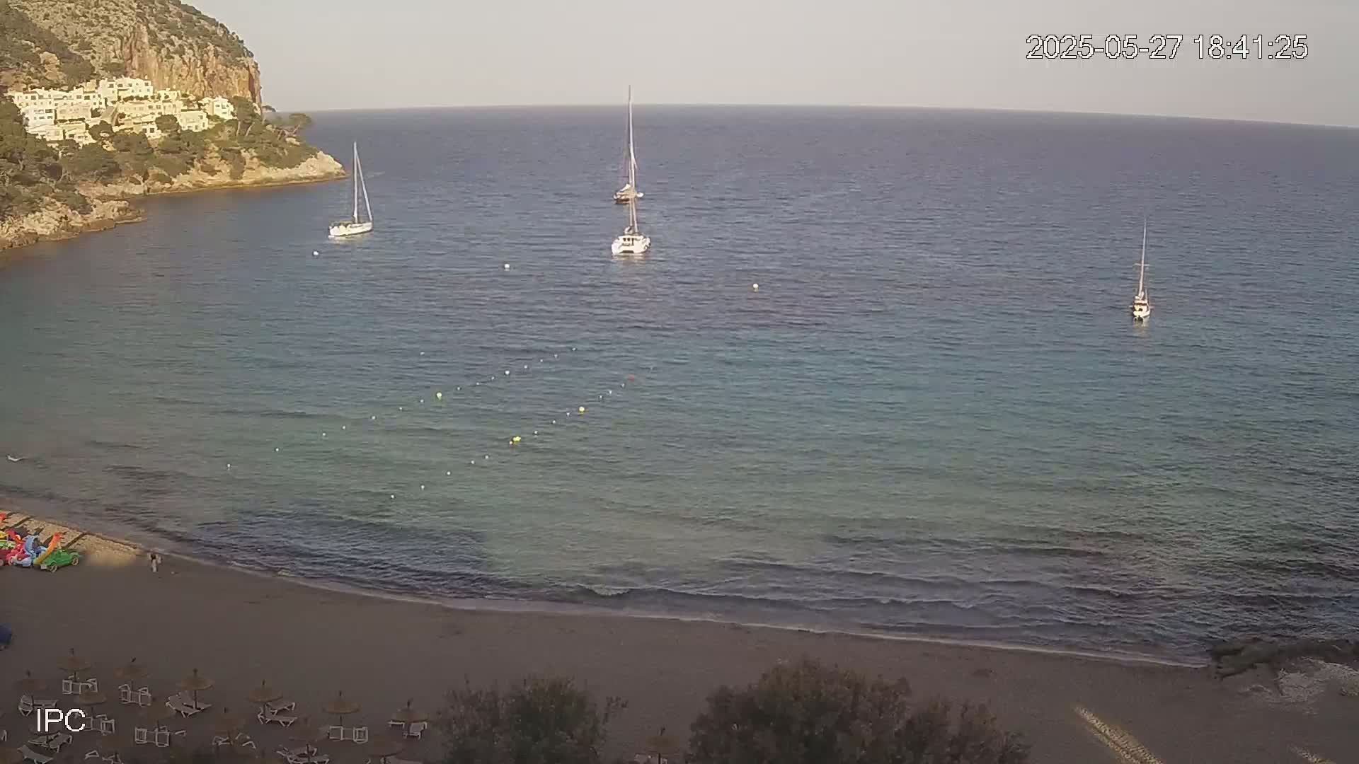 A sandy beach with calm, clear turquoise water and several sailboats anchored offshore, under a mostly clear sky.