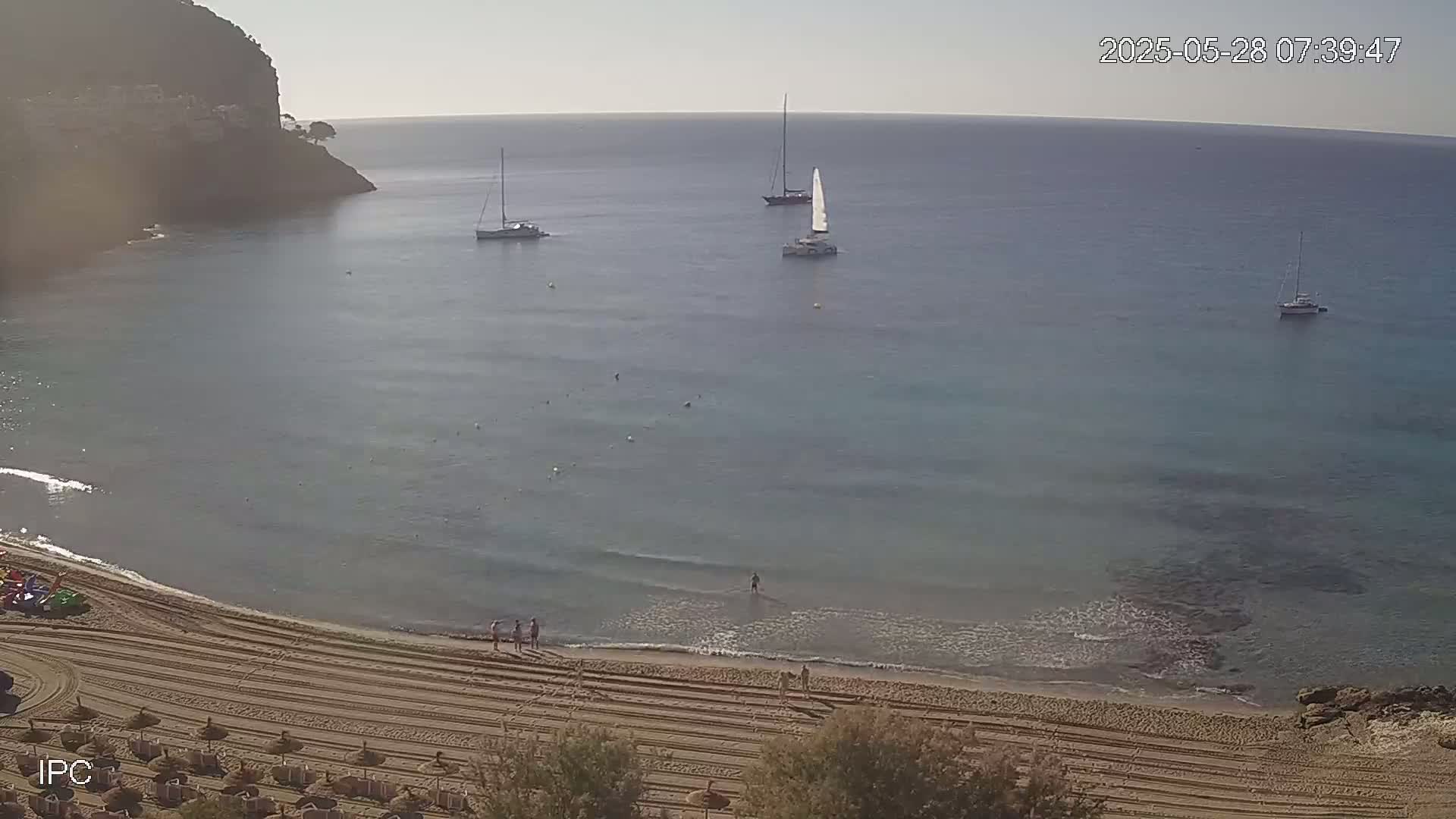 A sandy beach with several sailboats in calm, clear blue water, backed by a rocky cliff on a sunny day.