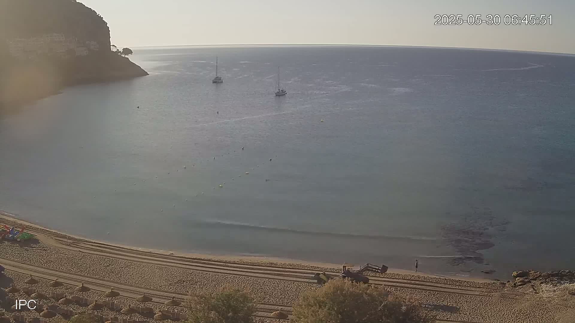 A calm, clear day reveals a sandy beach curving into a bay of tranquil, shallow water with two sailboats visible offshore, a cliff face rising behind the beach.