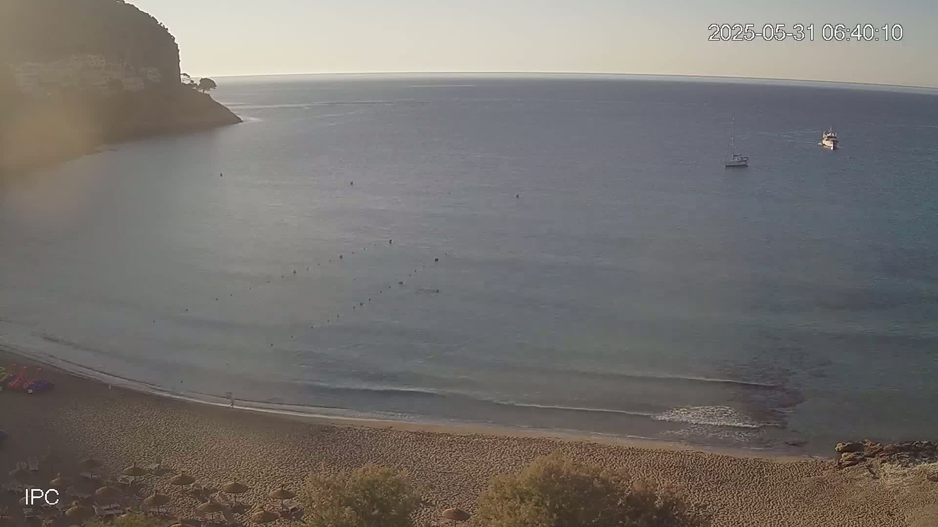 A sandy beach curves into a calm, light-blue sea under a clear morning sky, with two small boats visible on the water and a rocky cliff in the background.