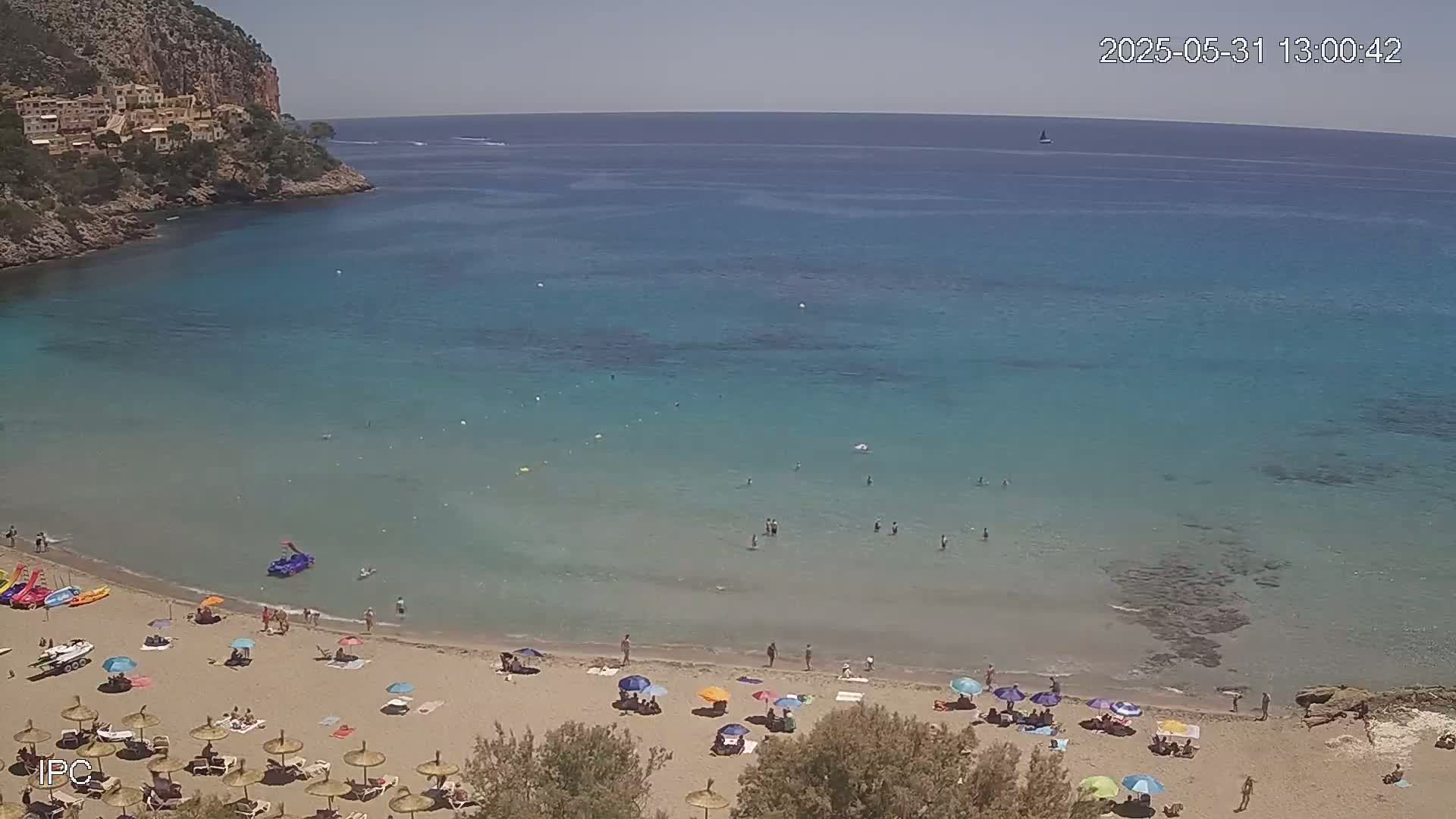 A sandy beach with numerous umbrellas and people is nestled in a calm, clear turquoise cove, backed by a rocky cliff face under a sunny sky.