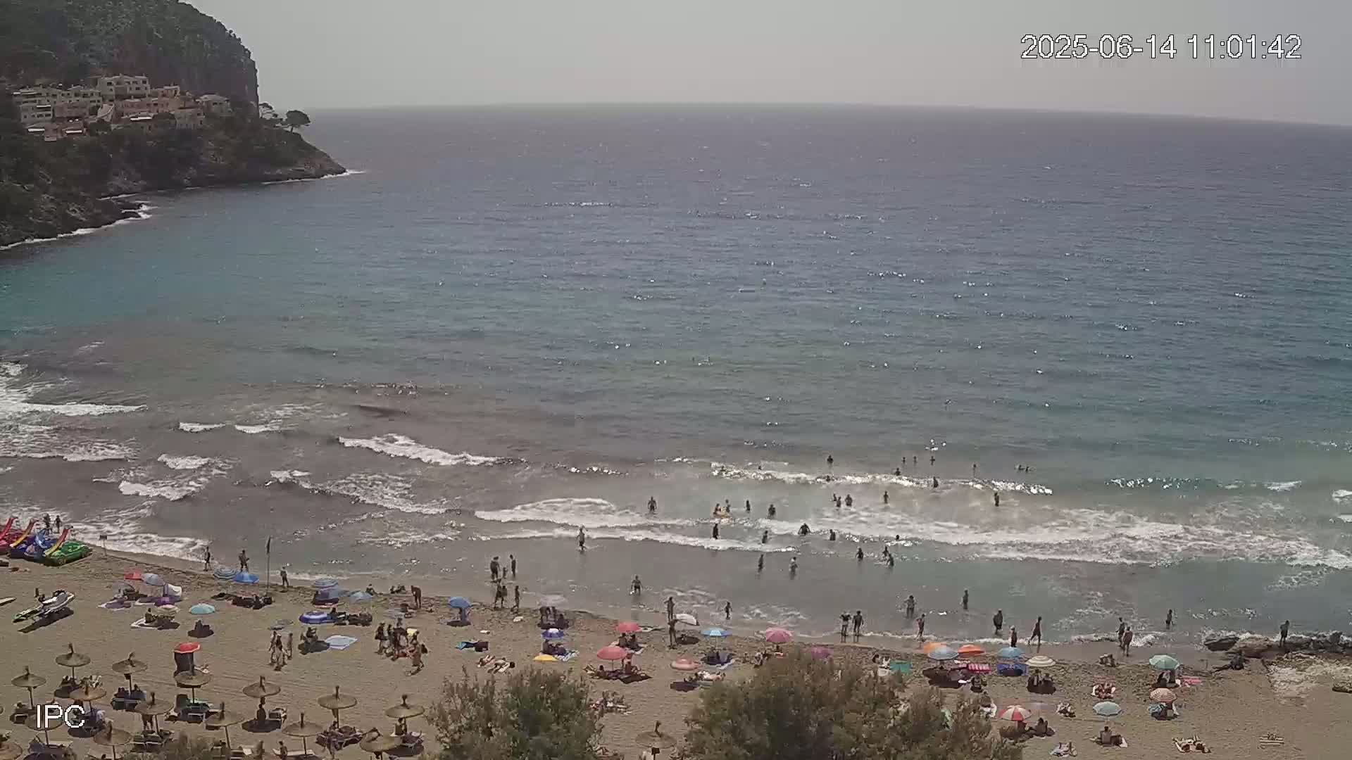 A sandy beach with many people under umbrellas and swimming in a calm sea near a rocky cliff on a sunny day.