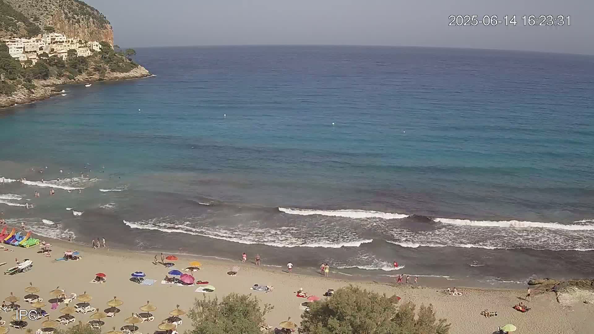 A sandy beach with people swimming and sunbathing under umbrellas, bordered by calm, turquoise water with small waves lapping the shore, backed by a rocky cliff with buildings.
