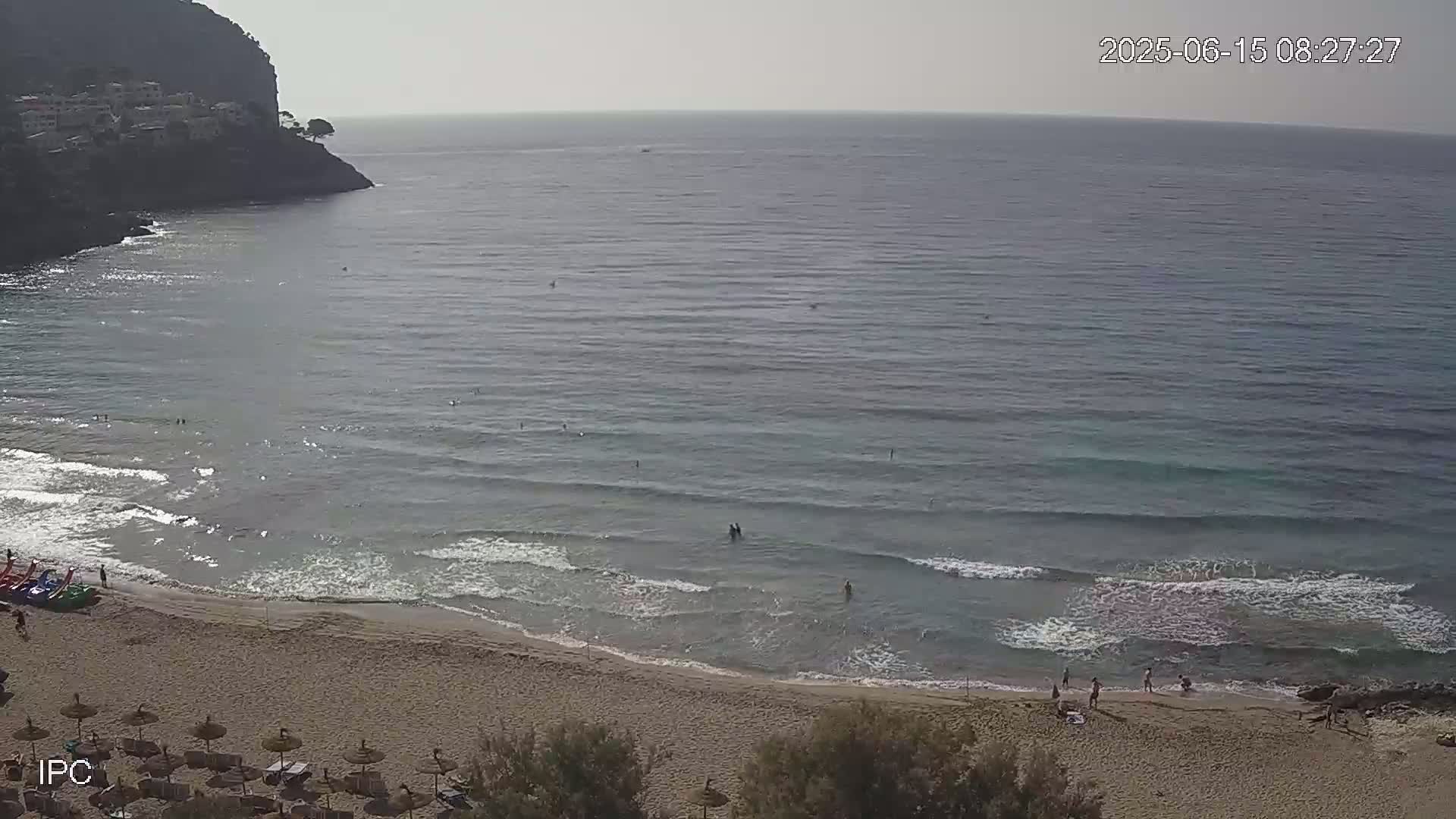 An aerial view shows a sandy beach with people swimming and sunbathing in calm, clear water under a hazy sky, backed by a rocky cliff.