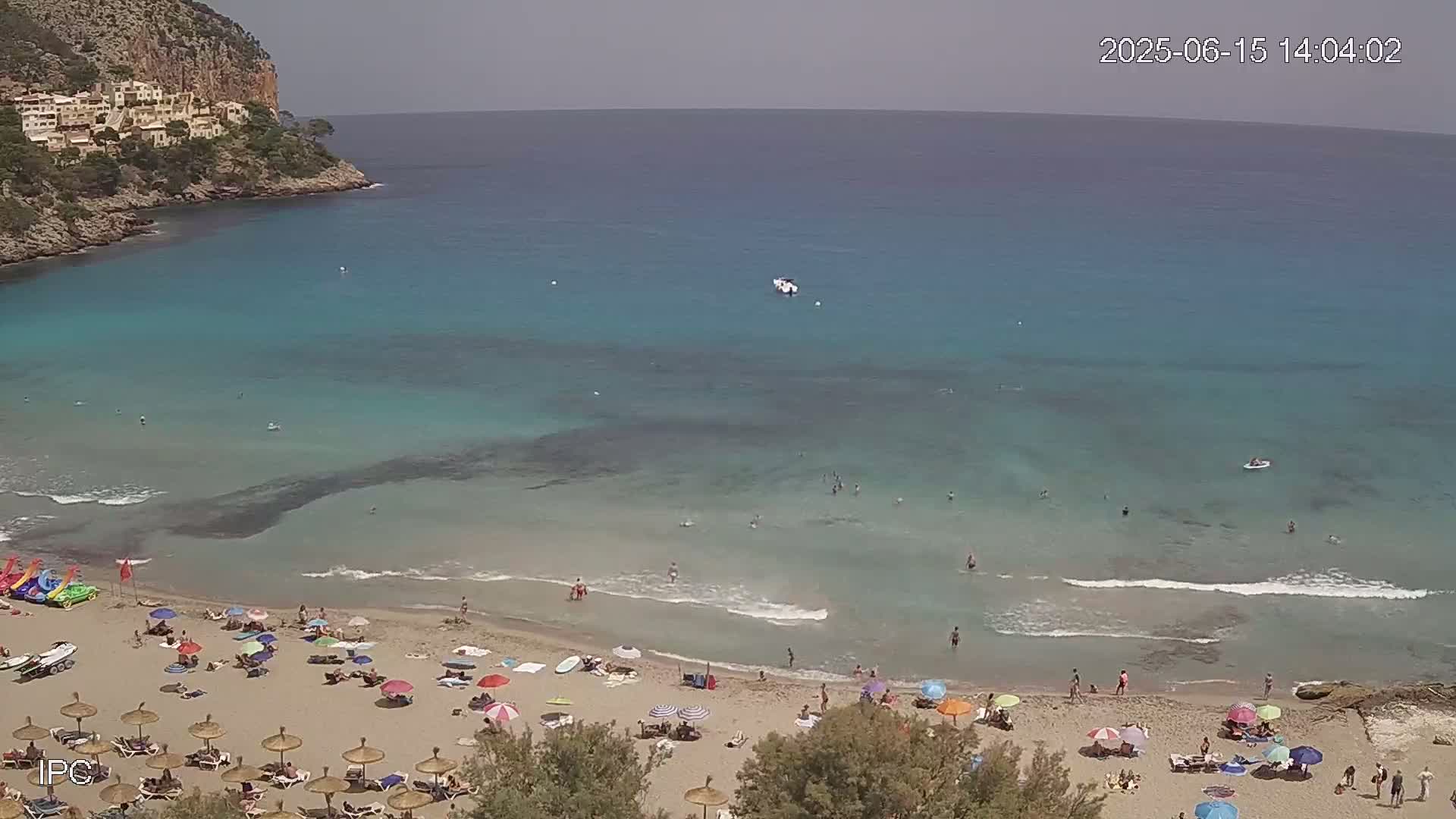 A sandy beach with many people and colorful umbrellas is nestled in a cove with clear turquoise water, under a mostly sunny sky.