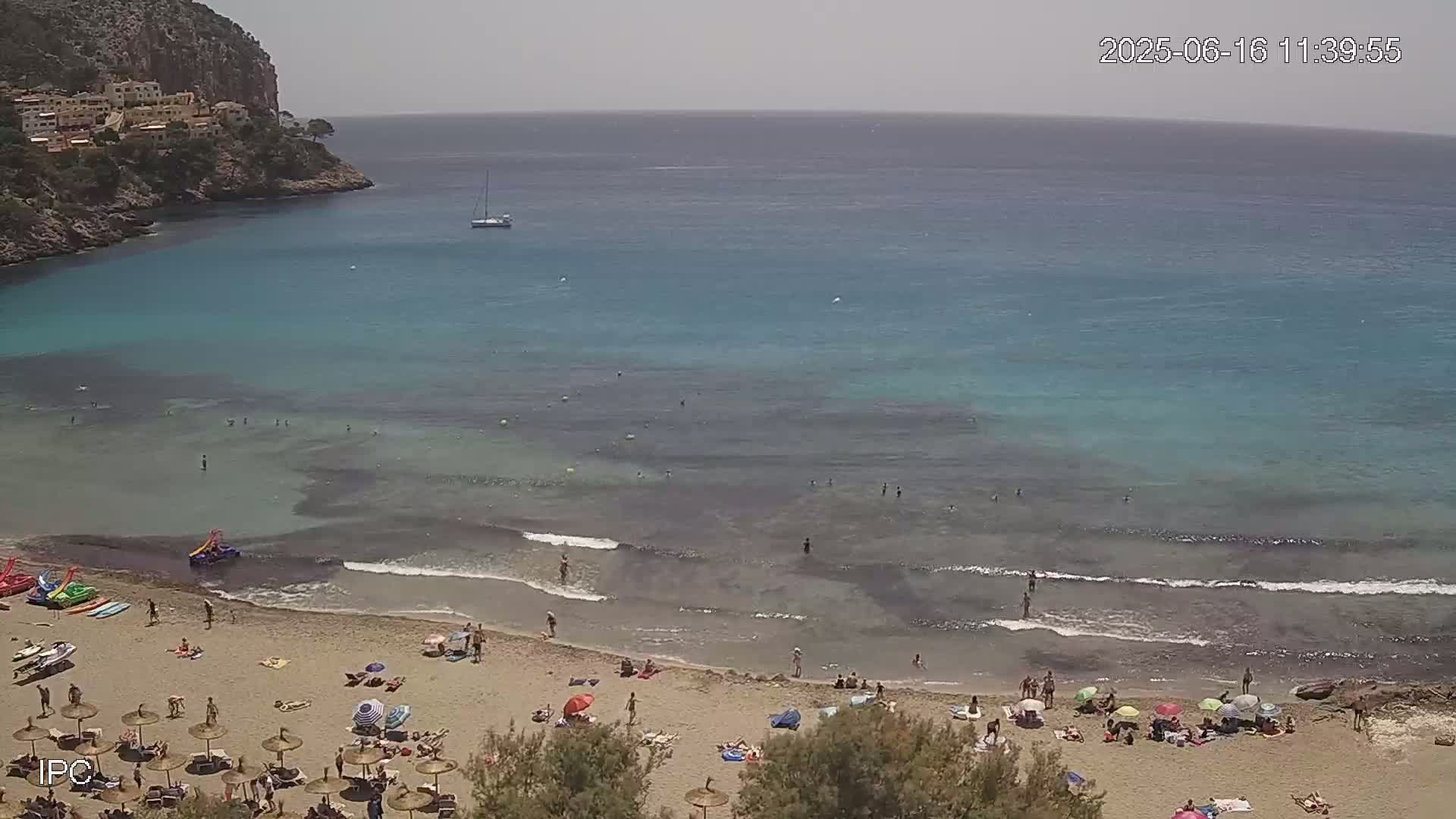 A sandy beach with clear, shallow turquoise water, populated by bathers and umbrellas under a mostly sunny sky, is nestled in a cove next to a rocky cliff and a small sailboat.