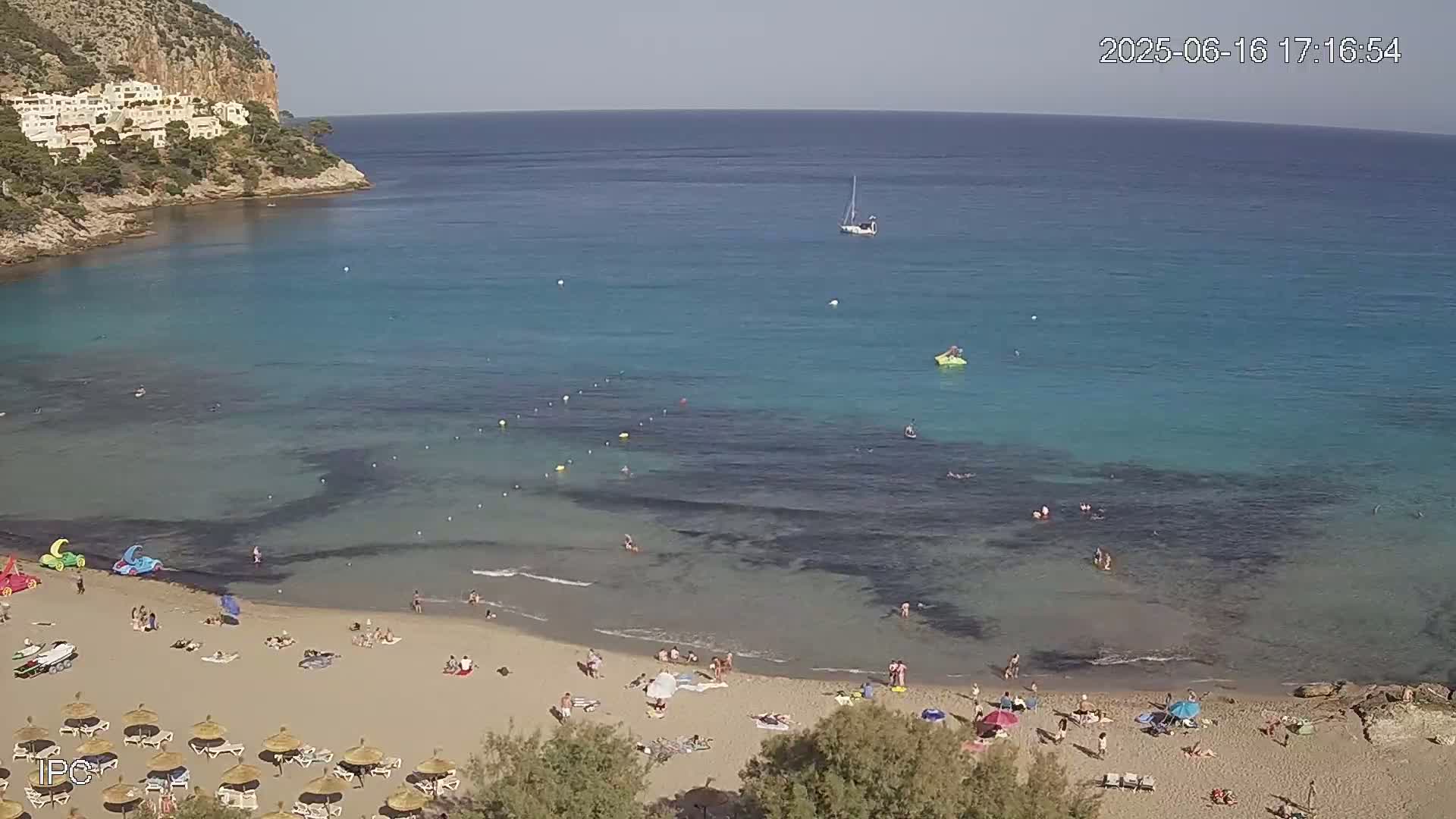 A sandy beach with many people on it is nestled in a cove of clear, turquoise water under a sunny sky, with a sailboat visible further out to sea.