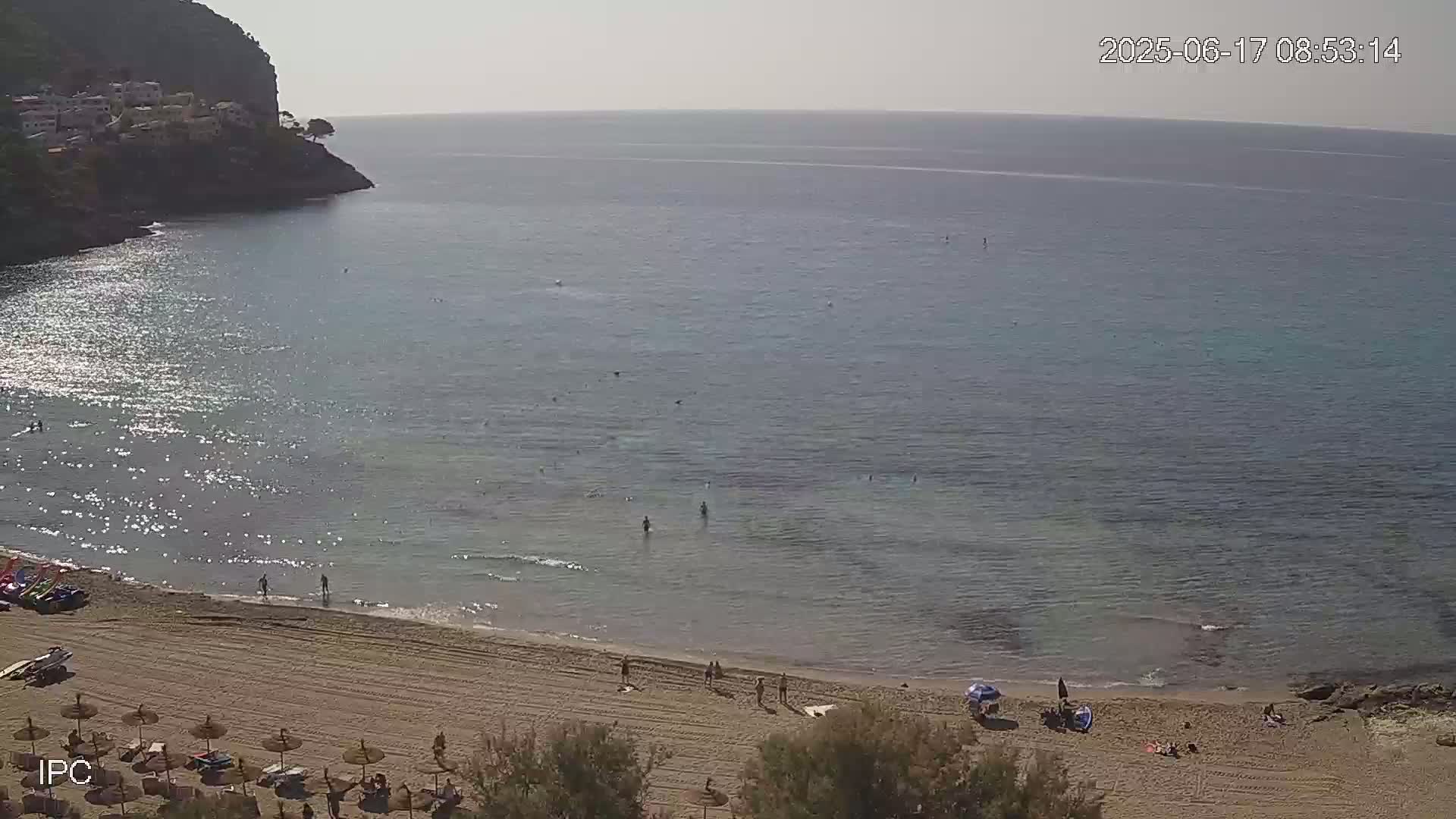 A sandy beach with scattered sun umbrellas and people, sits below a rocky cliff next to a calm, clear, sparkling sea on a sunny day.