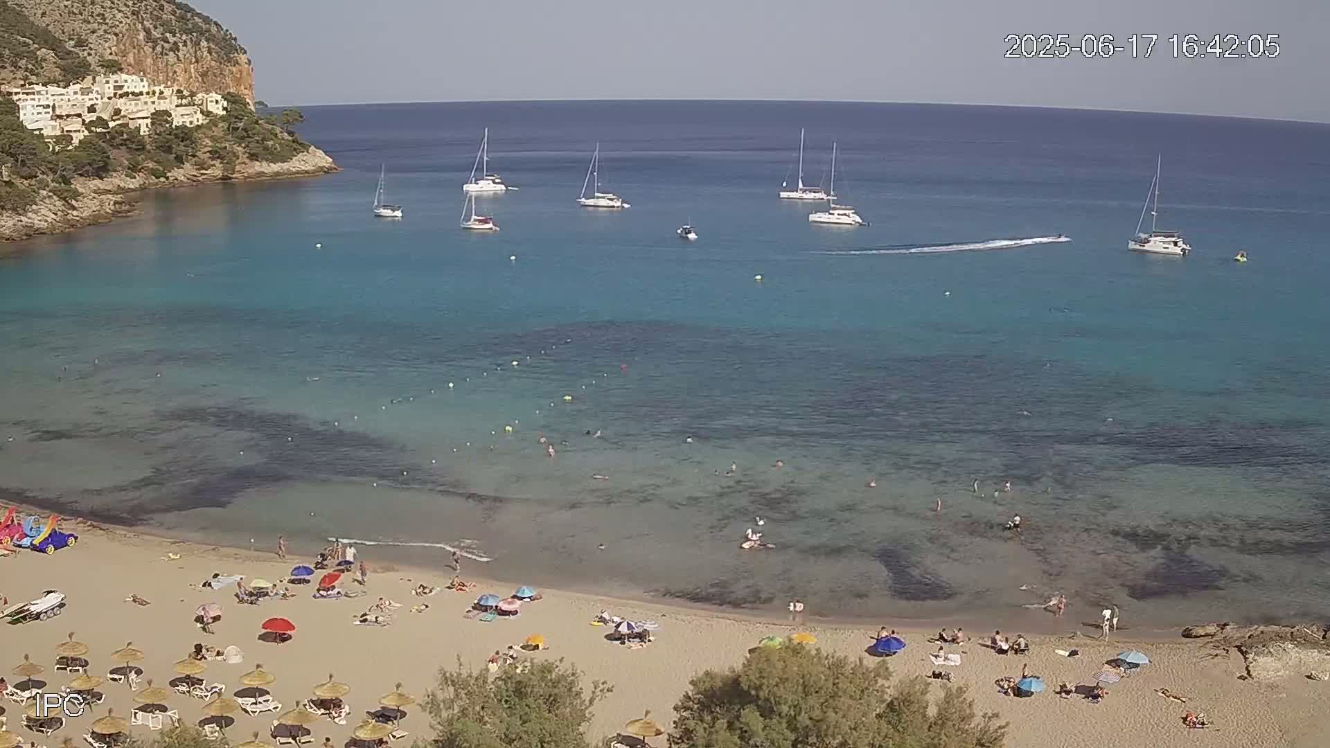 A sandy beach with numerous sunbathers and umbrellas is nestled in a calm, clear turquoise bay, surrounded by a rocky cliff and several sailboats under a sunny sky.
